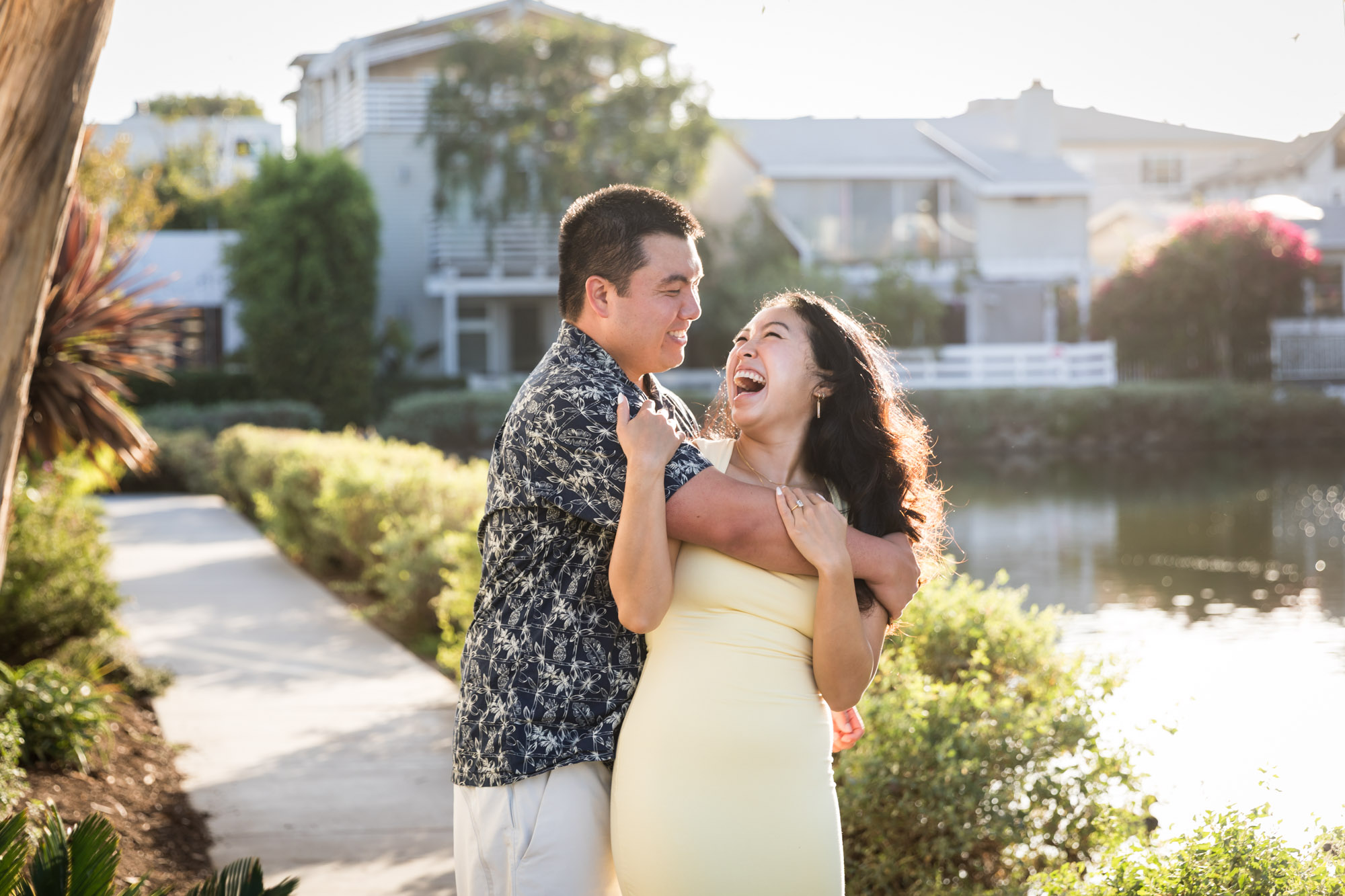 Man wearing blue and white patterned shirt hugging woman wearing sleeveless yellow dress from behind with canals in background during a Venice Canals engagement photo shoot