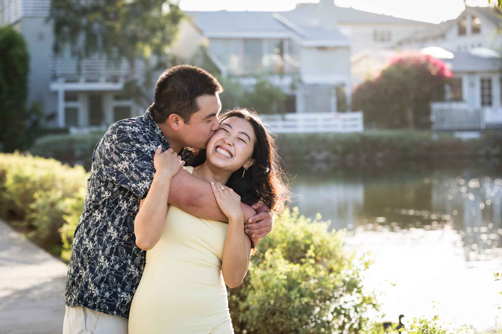 Man wearing blue and white patterned shirt kissing woman wearing sleeveless yellow dress from behind with canals in background during a Venice Canals engagement photo shoot