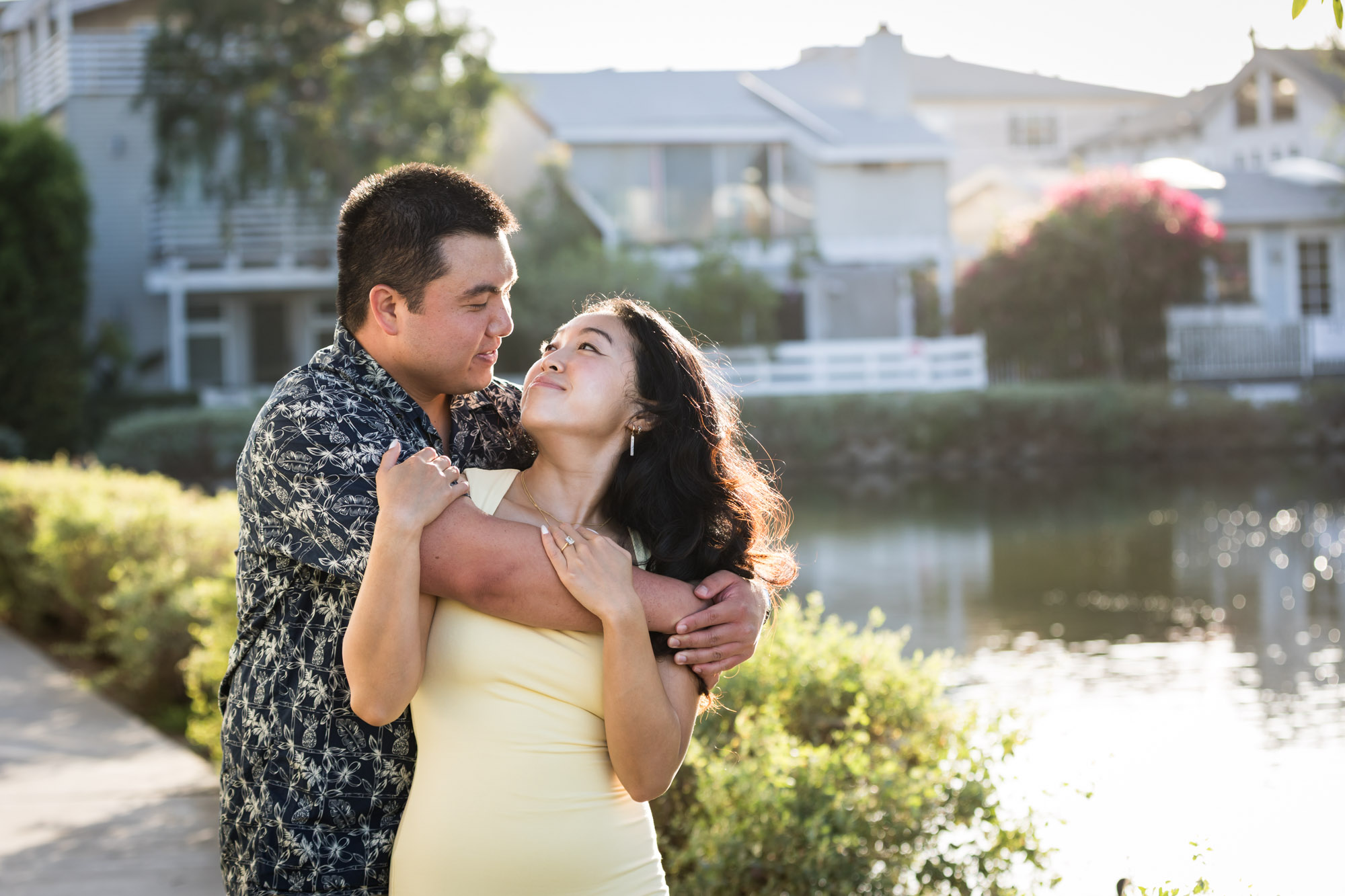 Man wearing blue and white patterned shirt hugging woman wearing sleeveless yellow dress from behind with canals in background during a Venice Canals engagement photo shoot