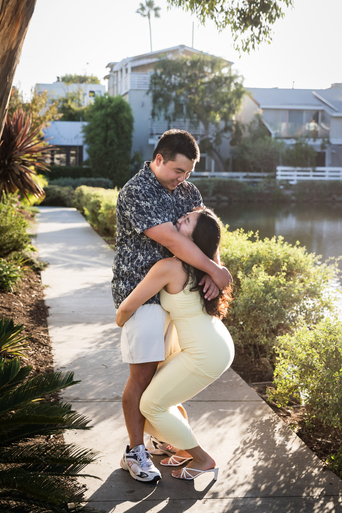 Woman wearing sleeveless yellow dress lifting man wearing blue and white patterned shirt in the air with canals in background during a Venice Canals engagement photo shoot