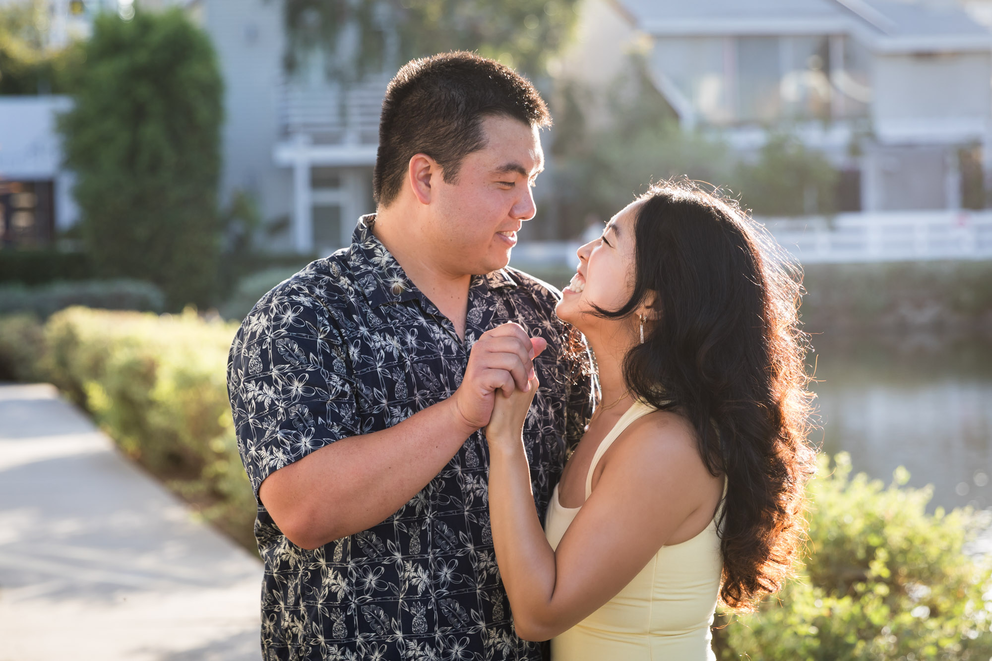Woman wearing sleeveless yellow dress holding hands with man wearing blue and white patterned shirt with canals in background during a Venice Canals engagement photo shoot