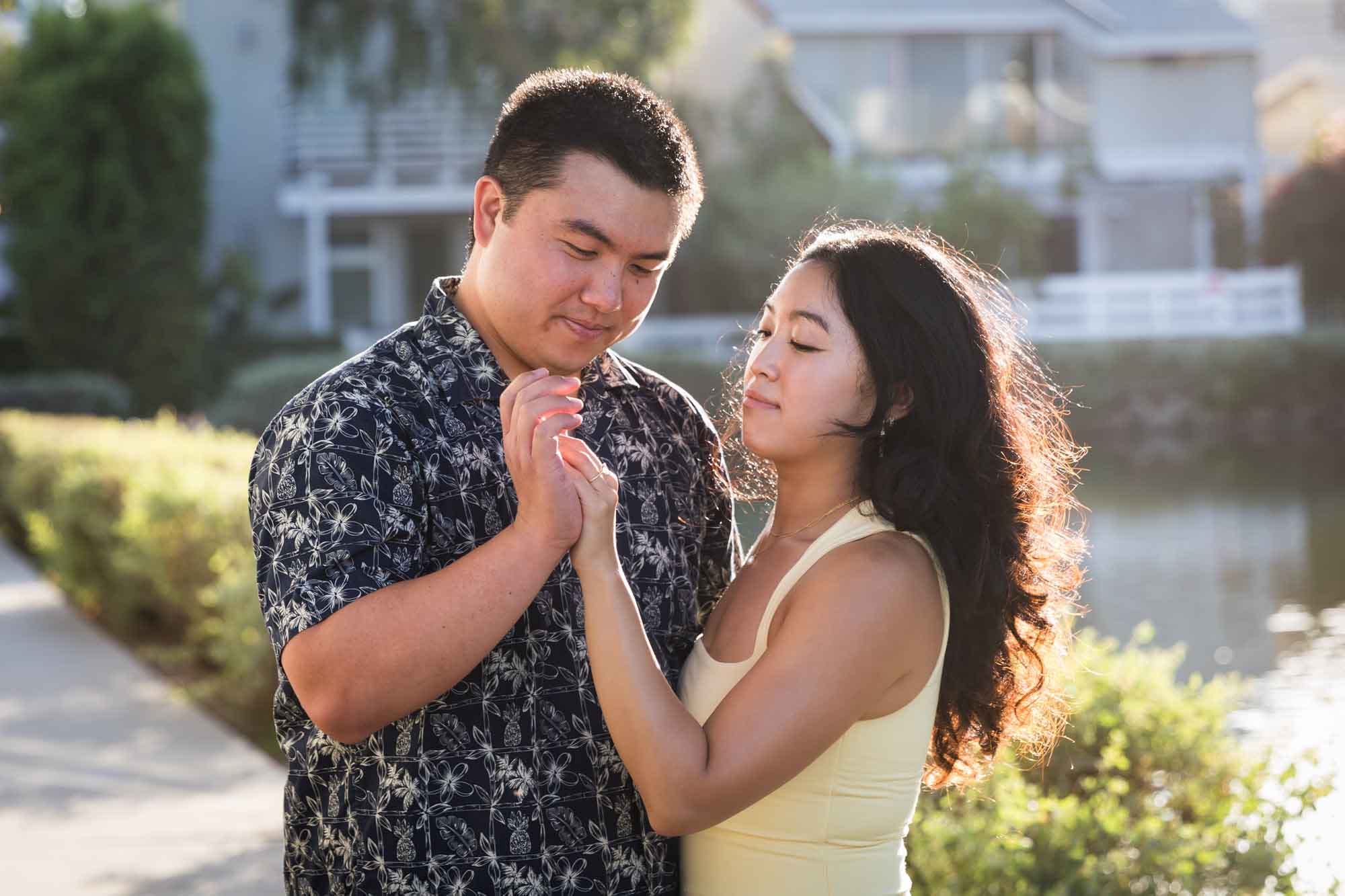 Woman wearing sleeveless yellow dress holding hands with man wearing blue and white patterned shirt with canals in background during a Venice Canals engagement photo shoot