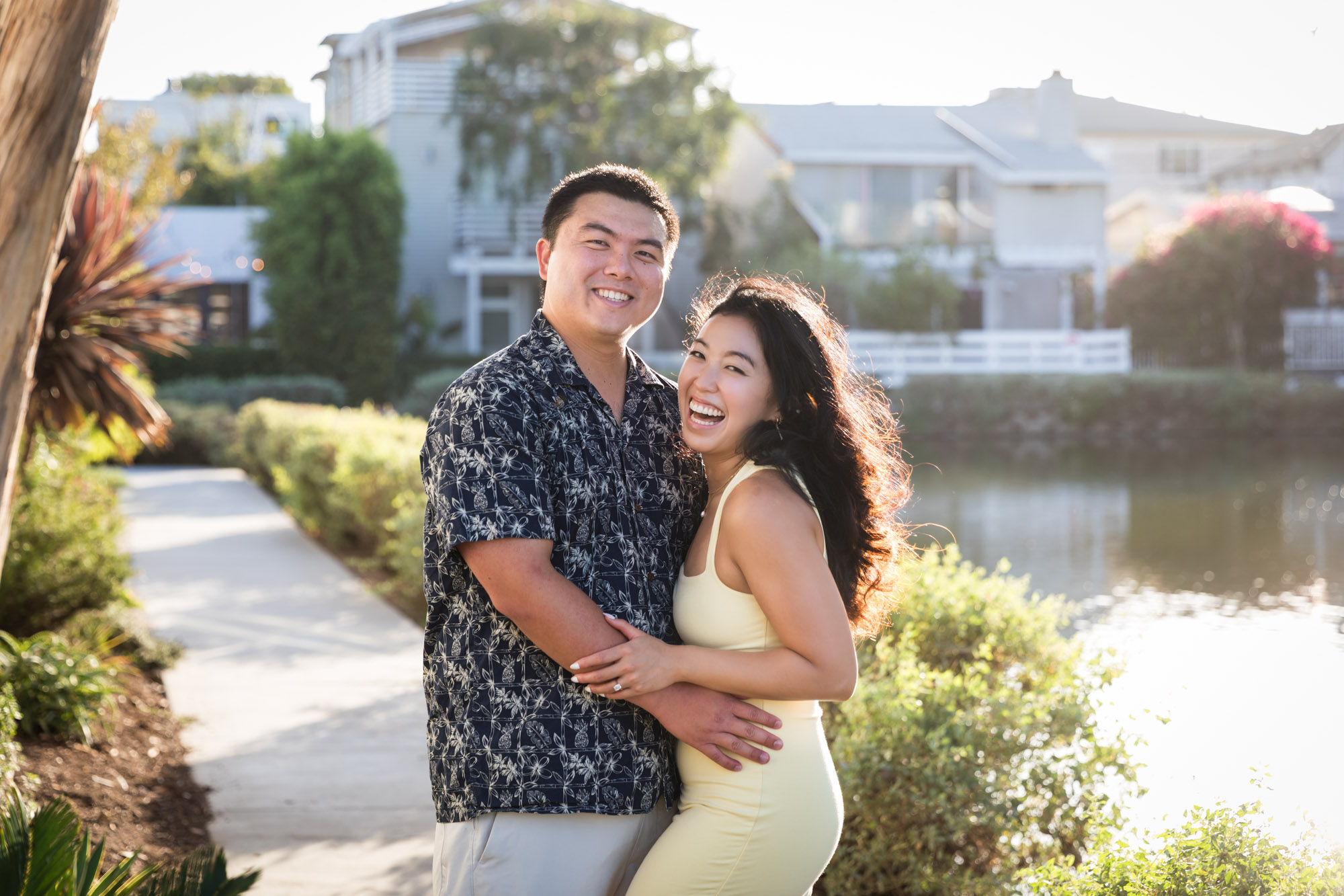 Woman wearing sleeveless yellow dress hugging man wearing blue and white patterned shirt with canals in background during a Venice Canals engagement photo shoot