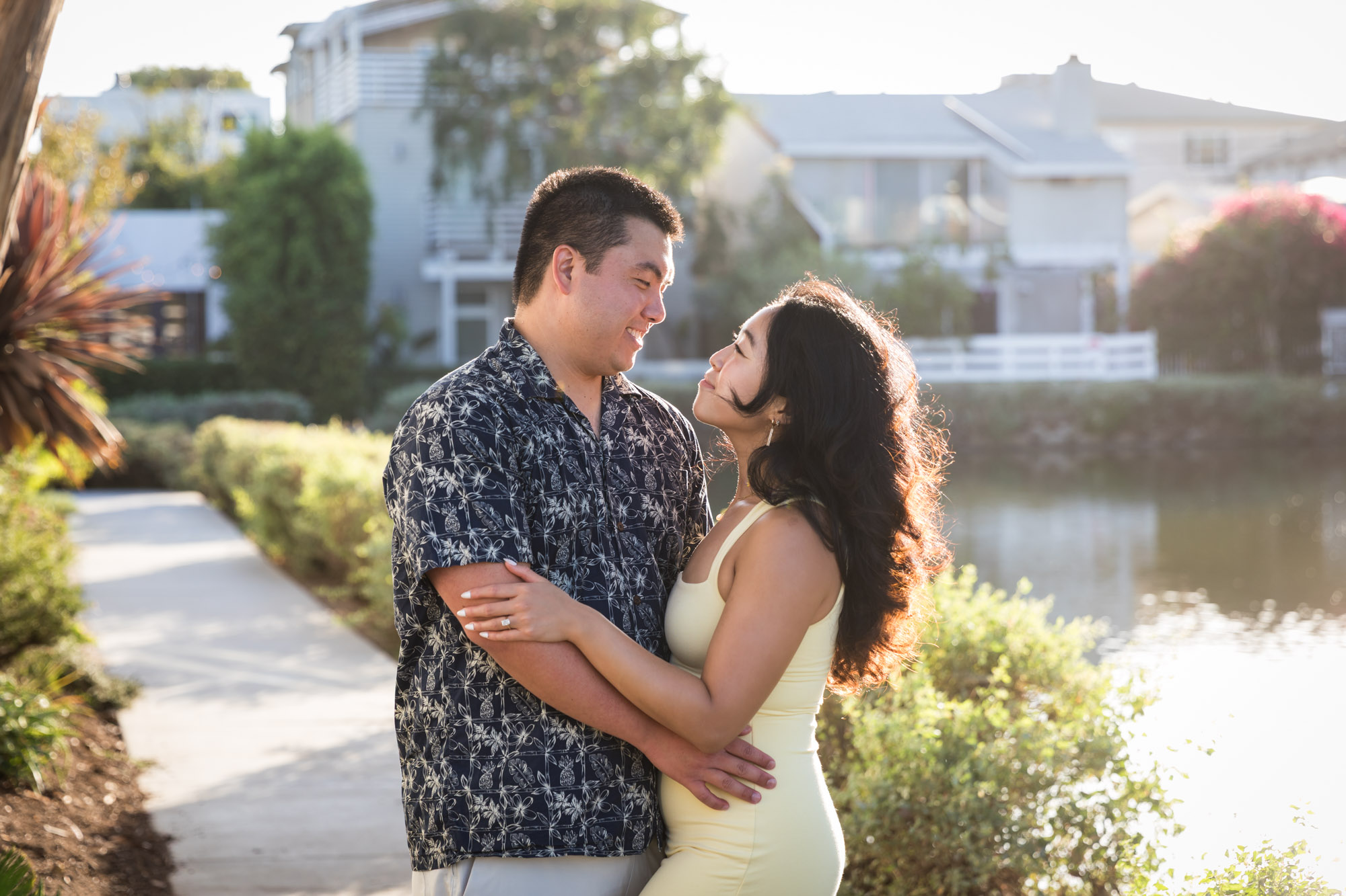 Woman wearing sleeveless yellow dress hugging man wearing blue and white patterned shirt with canals in background during a Venice Canals engagement photo shoot