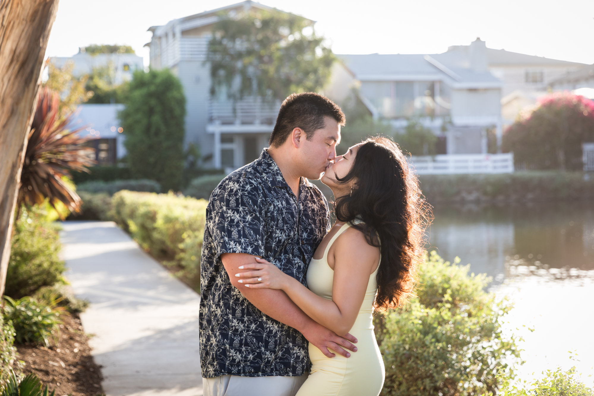 Woman wearing sleeveless yellow dress kissing man wearing blue and white patterned shirt with canals in background during a Venice Canals engagement photo shoot