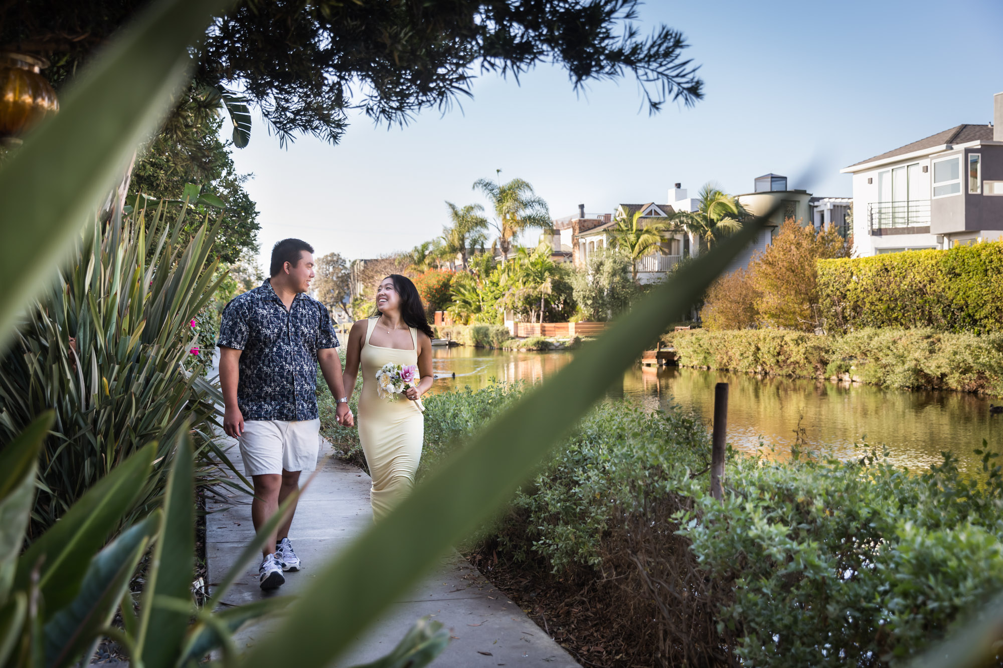 Woman wearing long yellow dress walking hand-in-hand with man wearing blue and white patterned shirt along pathway beside canal with house in background during a Venice Canals engagement photo shoot