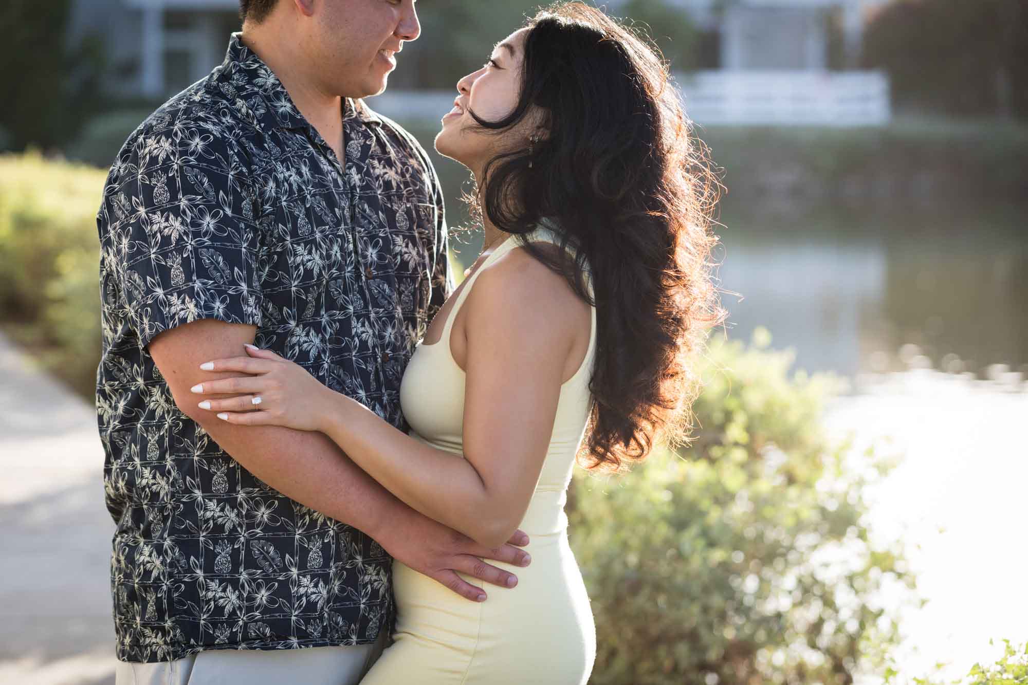 Woman wearing sleeveless yellow dress hugging man wearing blue and white patterned shirt with canals in background during a Venice Canals engagement photo shoot