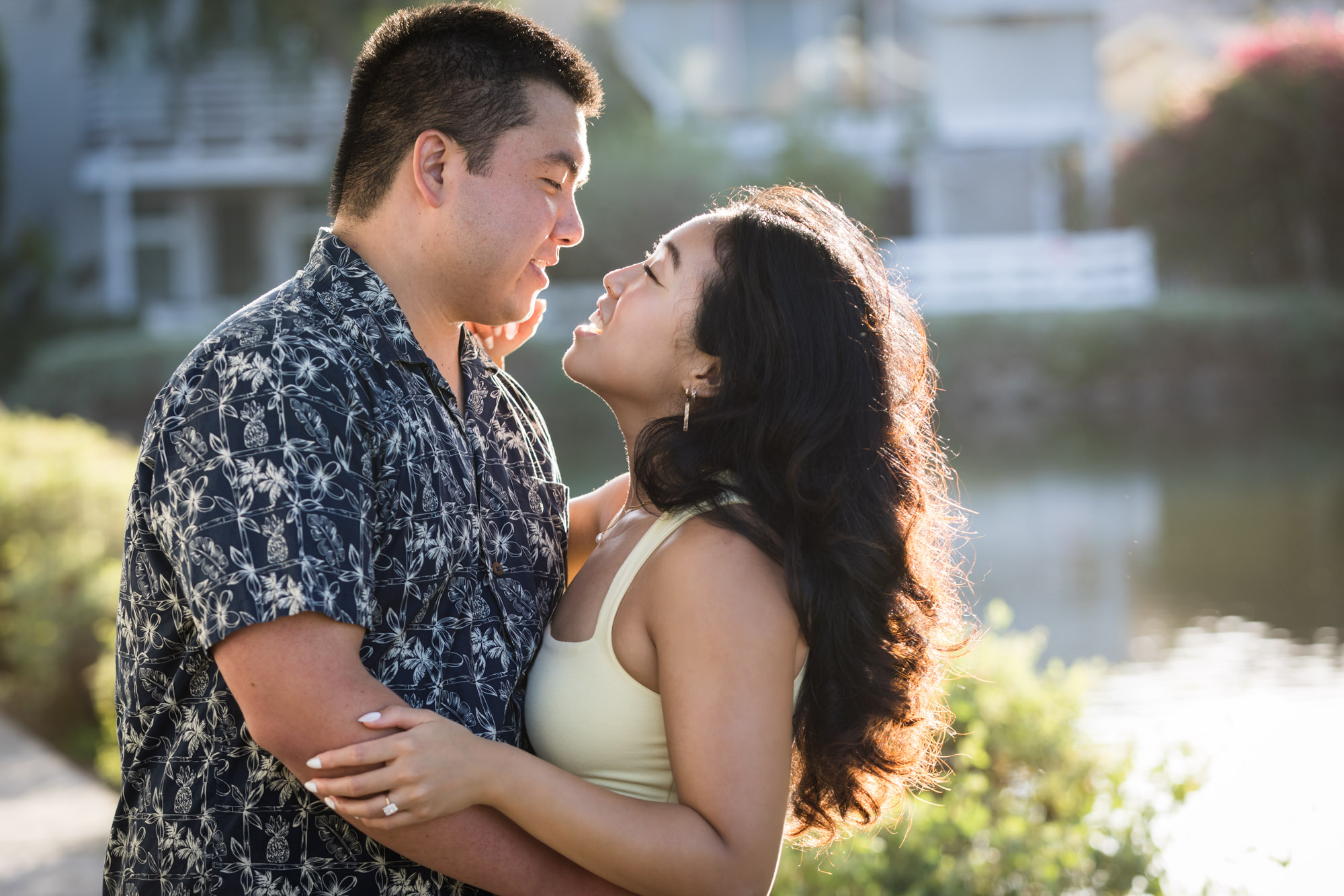 Woman wearing sleeveless yellow dress hugging man wearing blue and white patterned shirt with canals in background during a Venice Canals engagement photo shoot