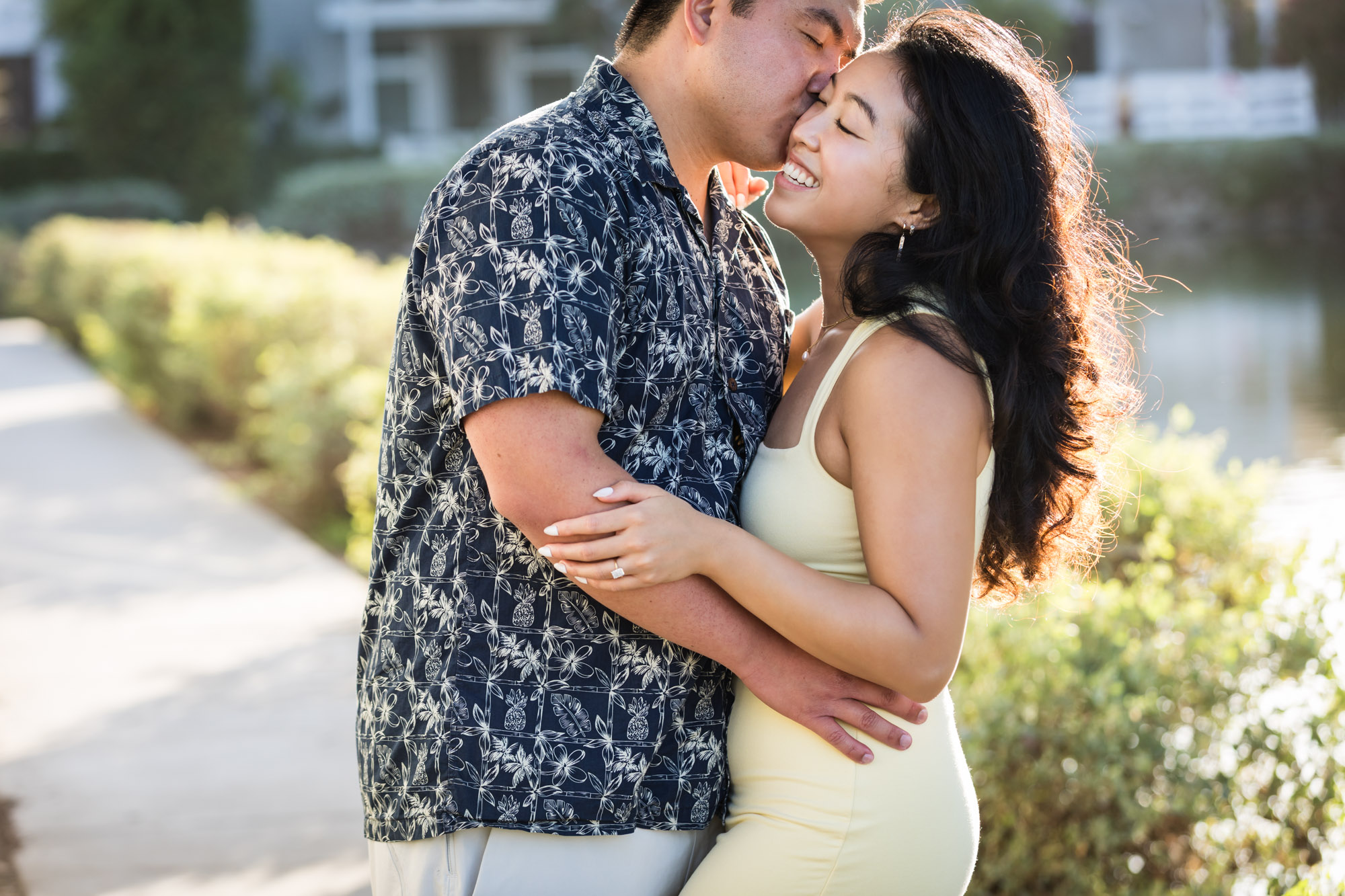 Woman wearing sleeveless yellow dress hugging man wearing blue and white patterned shirt with canals in background during a Venice Canals engagement photo shoot