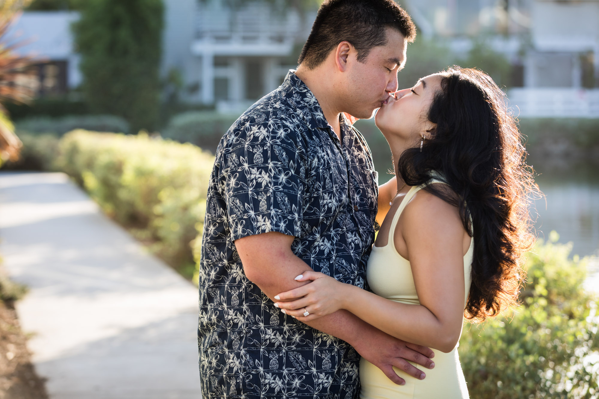 Woman wearing sleeveless yellow dress hugging man wearing blue and white patterned shirt with canals in background during a Venice Canals engagement photo shoot