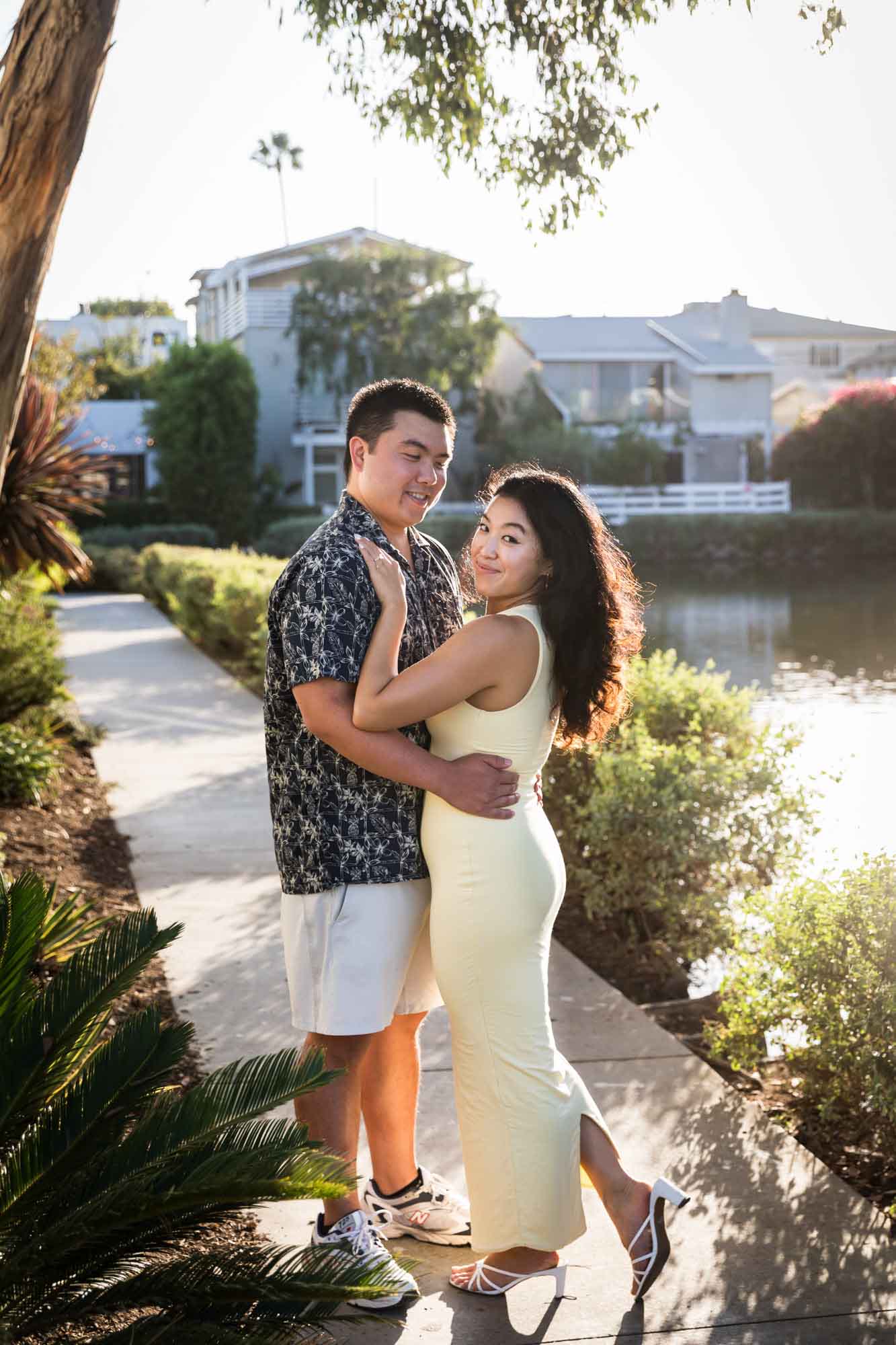 Woman wearing sleeveless yellow dress hugging man wearing blue and white patterned shirt with canals in background during a Venice Canals engagement photo shoot
