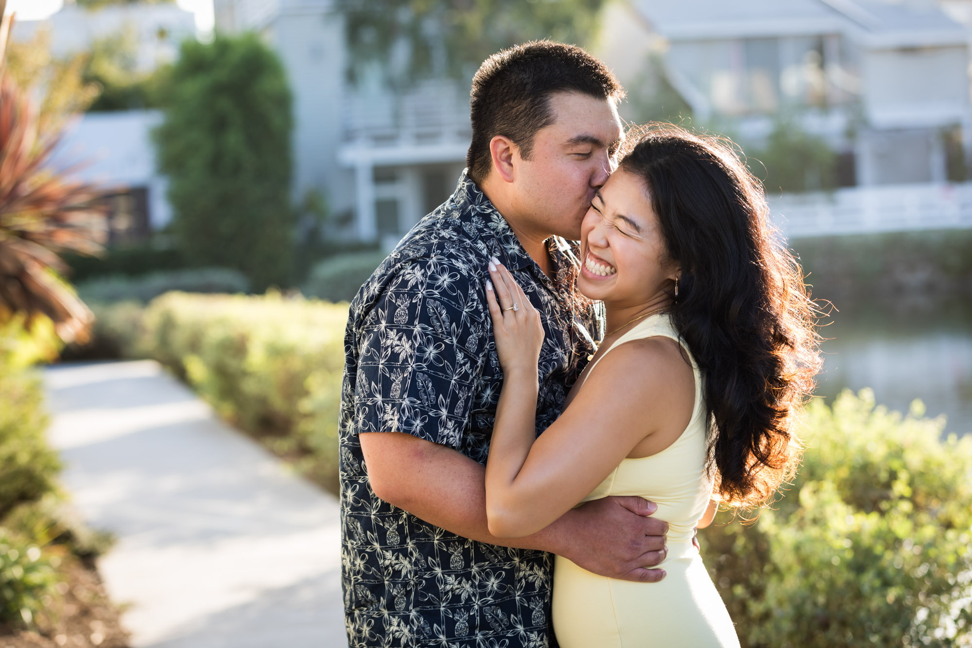 Woman wearing sleeveless yellow dress kissing man wearing blue and white patterned shirt with canals in background during a Venice Canals engagement photo shoot