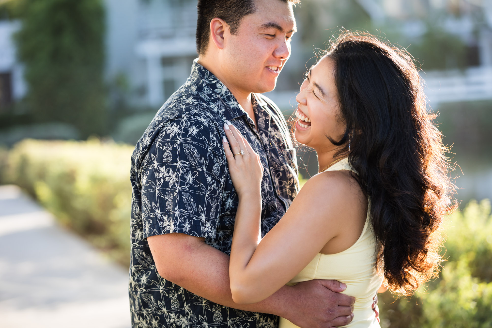 Woman wearing sleeveless yellow dress hugging man wearing blue and white patterned shirt with canals in background during a Venice Canals engagement photo shoot