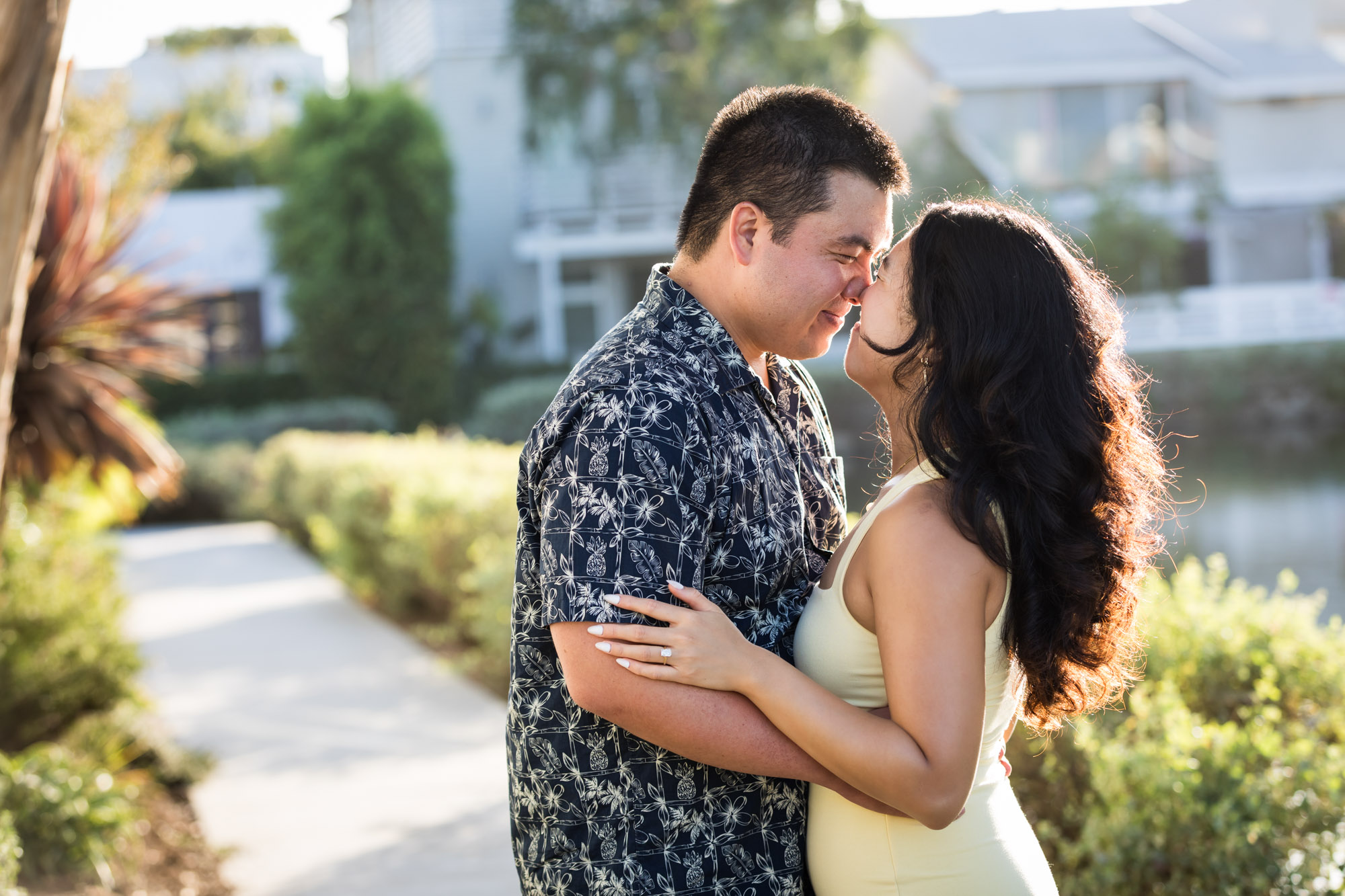 Woman wearing sleeveless yellow dress rubbing noses with man wearing blue and white patterned shirt with canals in background during a Venice Canals engagement photo shoot