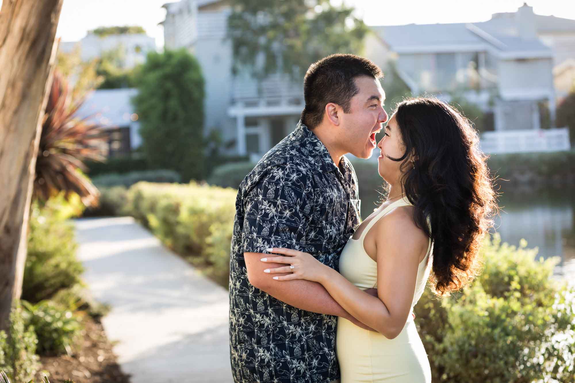 Woman wearing sleeveless yellow dress avoiding bite of man wearing blue and white patterned shirt with canals in background during a Venice Canals engagement photo shoot