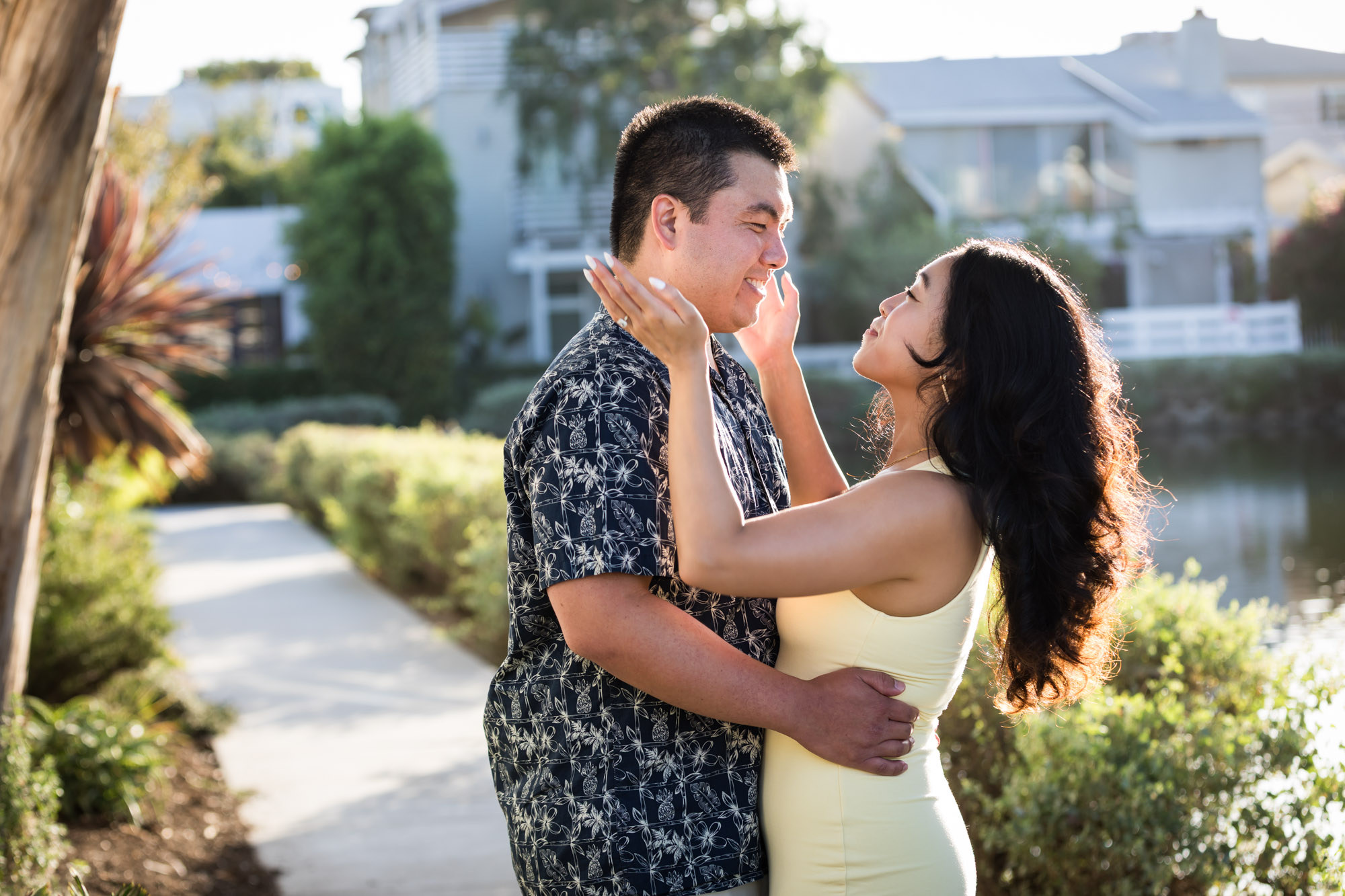 Woman wearing sleeveless yellow dress about to put hands around face of man wearing blue and white patterned shirt with canals in background during a Venice Canals engagement photo shoot