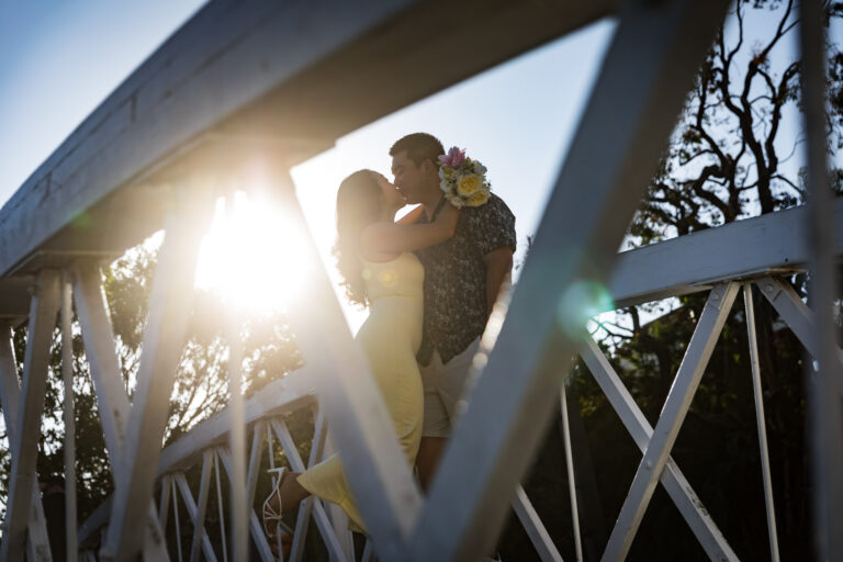 Woman wearing long yellow dress kissing man wearing blue and white shirt as seen in silhouette through the railing of a bridge during a Venice Canals engagement photo shoot