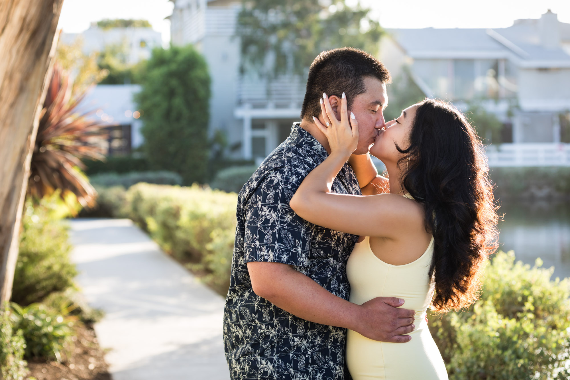 Woman wearing sleeveless yellow dress kissing man wearing blue and white patterned shirt with canals in background during a Venice Canals engagement photo shoot