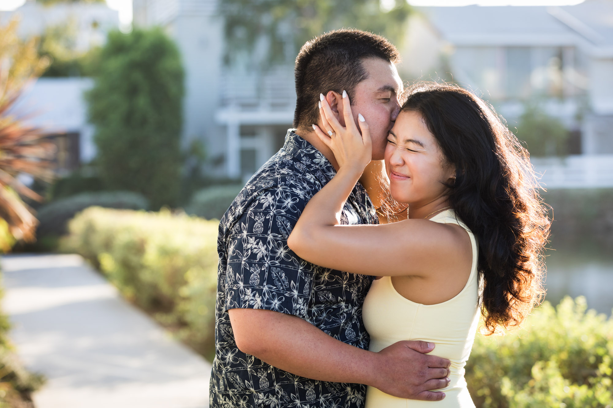 Woman wearing sleeveless yellow dress kissing man wearing blue and white patterned shirt with canals in background during a Venice Canals engagement photo shoot
