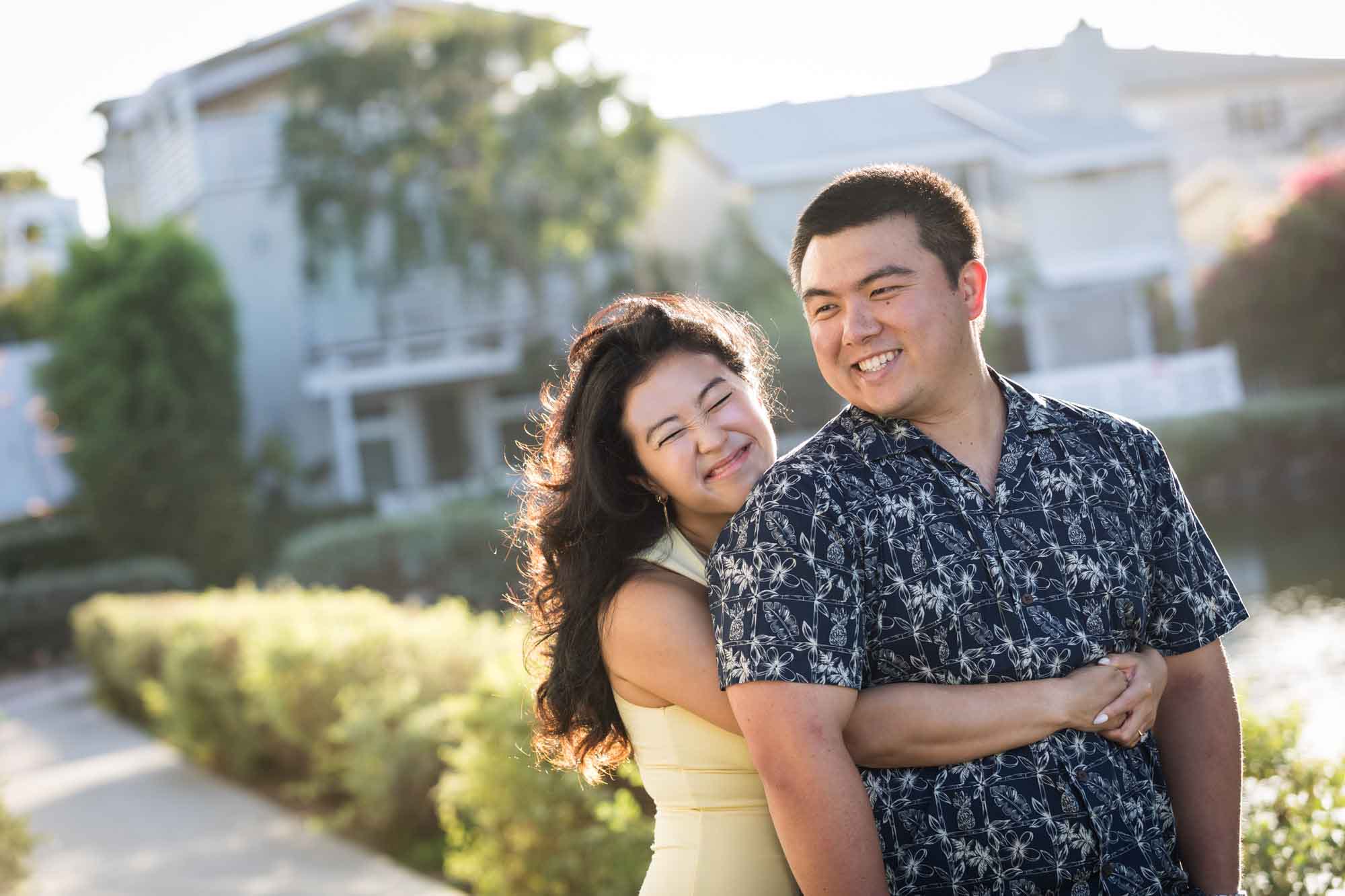 Woman wearing sleeveless yellow dress hugging man wearing blue and white patterned shirt with canals in background during a Venice Canals engagement photo shoot