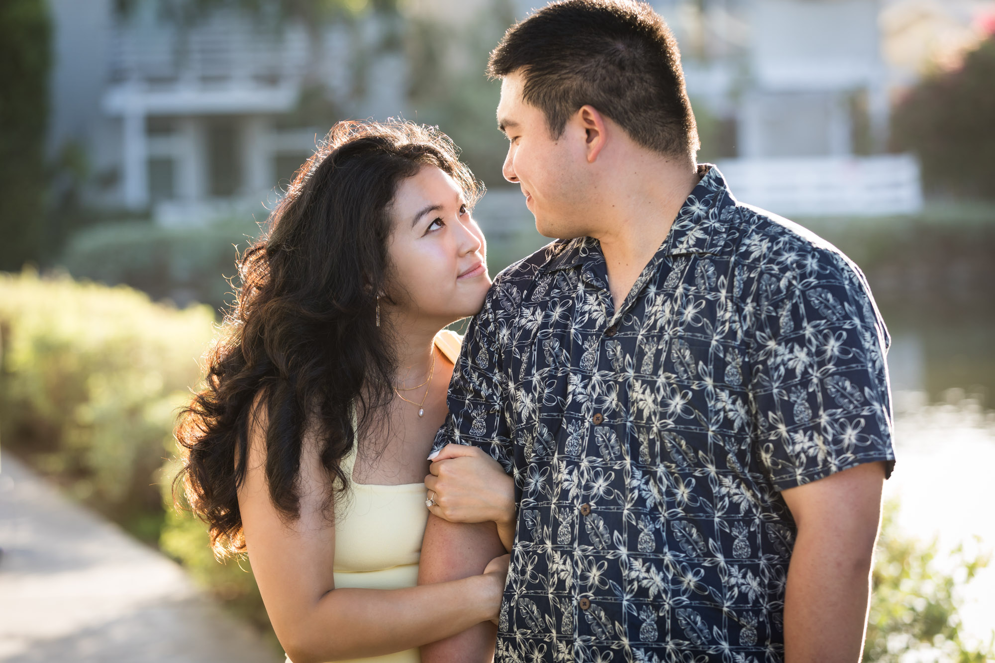 Woman wearing sleeveless yellow dress looking at man wearing blue and white patterned shirt with canals in background during a Venice Canals engagement photo shoot