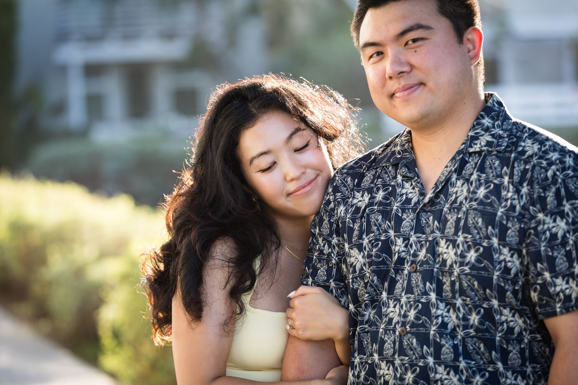 Woman wearing sleeveless yellow dress hugging man wearing blue and white patterned shirt with canals in background during a Venice Canals engagement photo shoot
