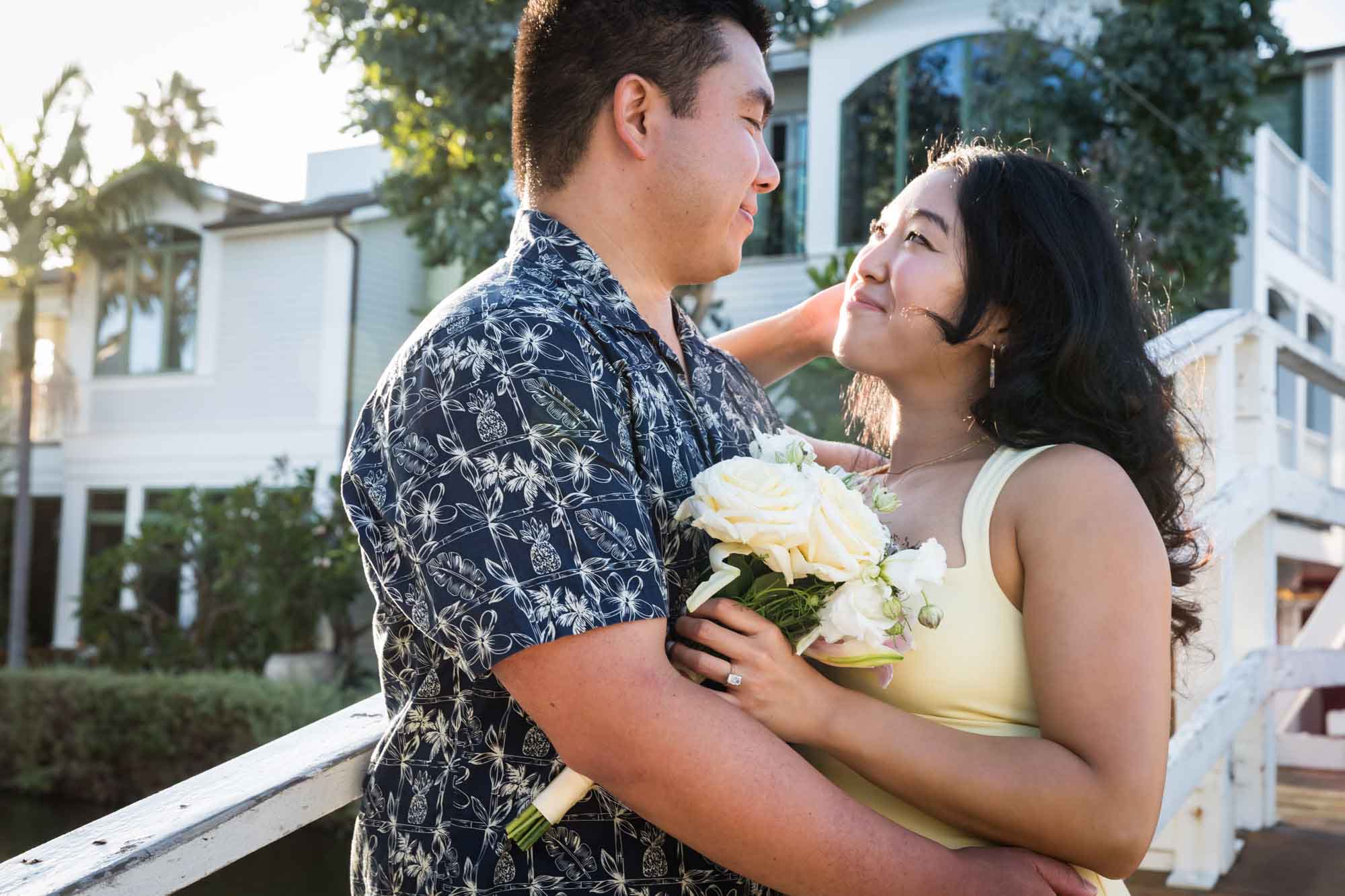 Woman wearing sleeveless yellow dress and man wearing blue and white patterned shirt looking at each other with canals in background during a Venice Canals engagement photo shoot