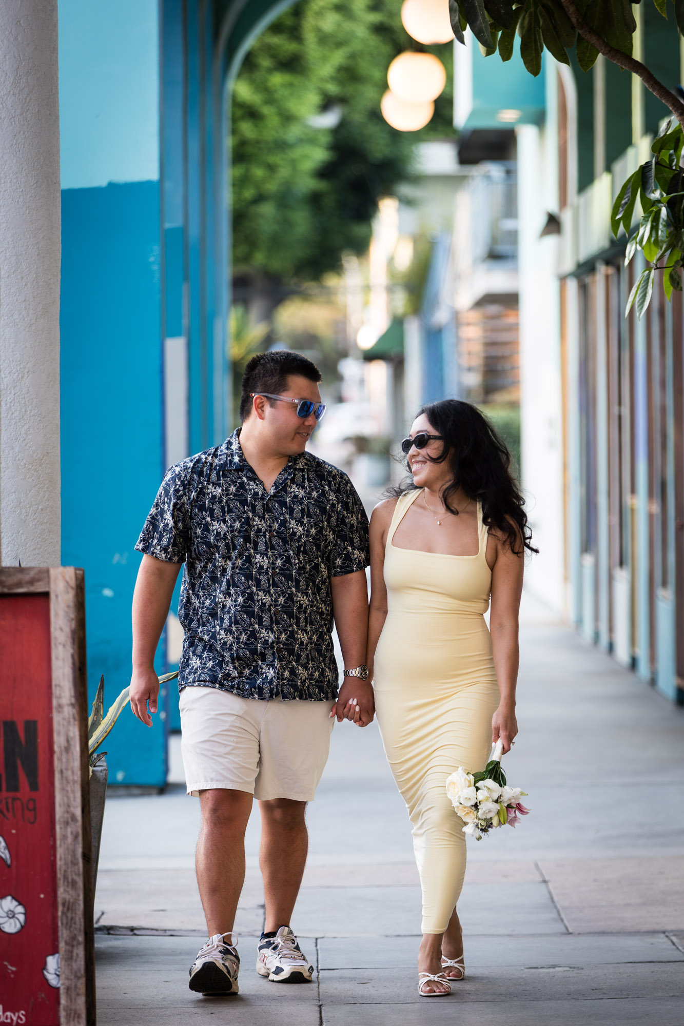 Man wearing blue and white patterned shirt and sunglasses and woman wearing sleeveless yellow dress and sunglasses walking down colonnade pathway during a Venice Canals engagement photo shoot
