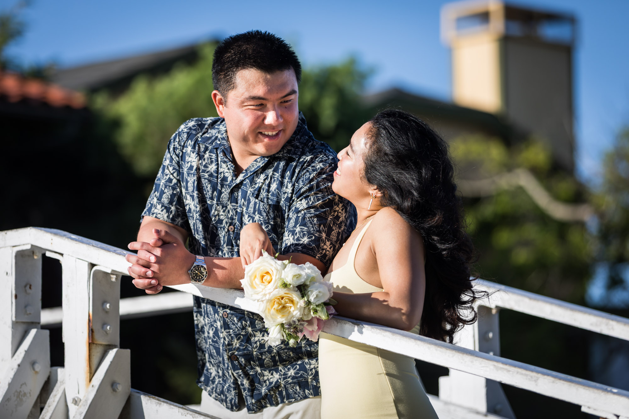 Woman wearing long yellow dress holding flowers on white bridge next to man wearing blue and white patterned shirt during a Venice Canals engagement photo shoot