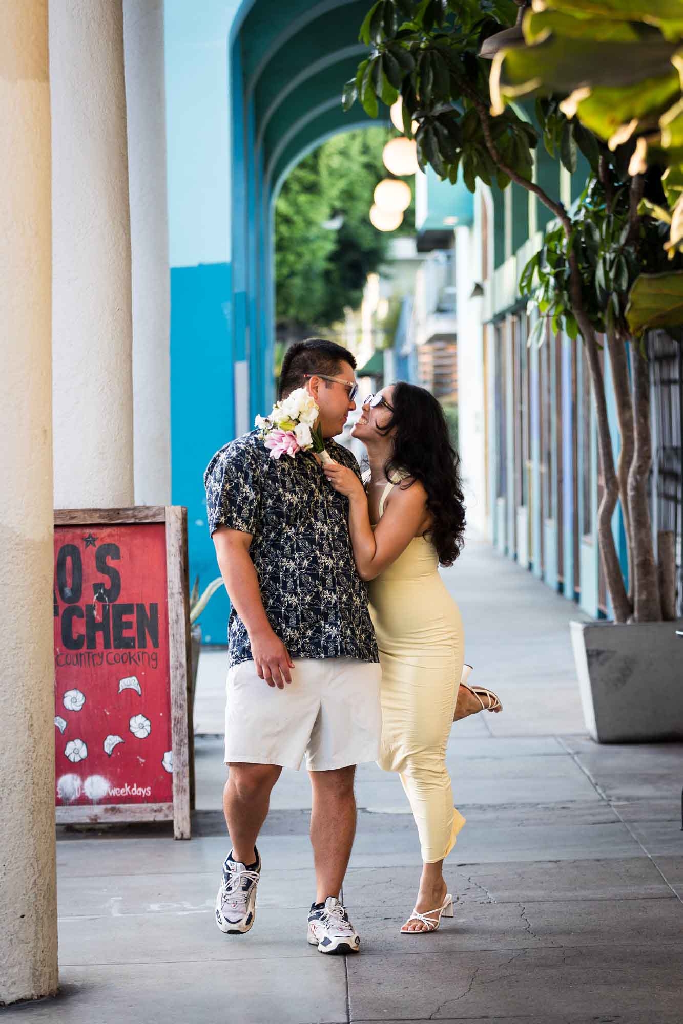 Man wearing blue and white patterned shirt and sunglasses and woman wearing sleeveless yellow dress and sunglasses walking down colonnade pathway during a Venice Canals engagement photo shoot