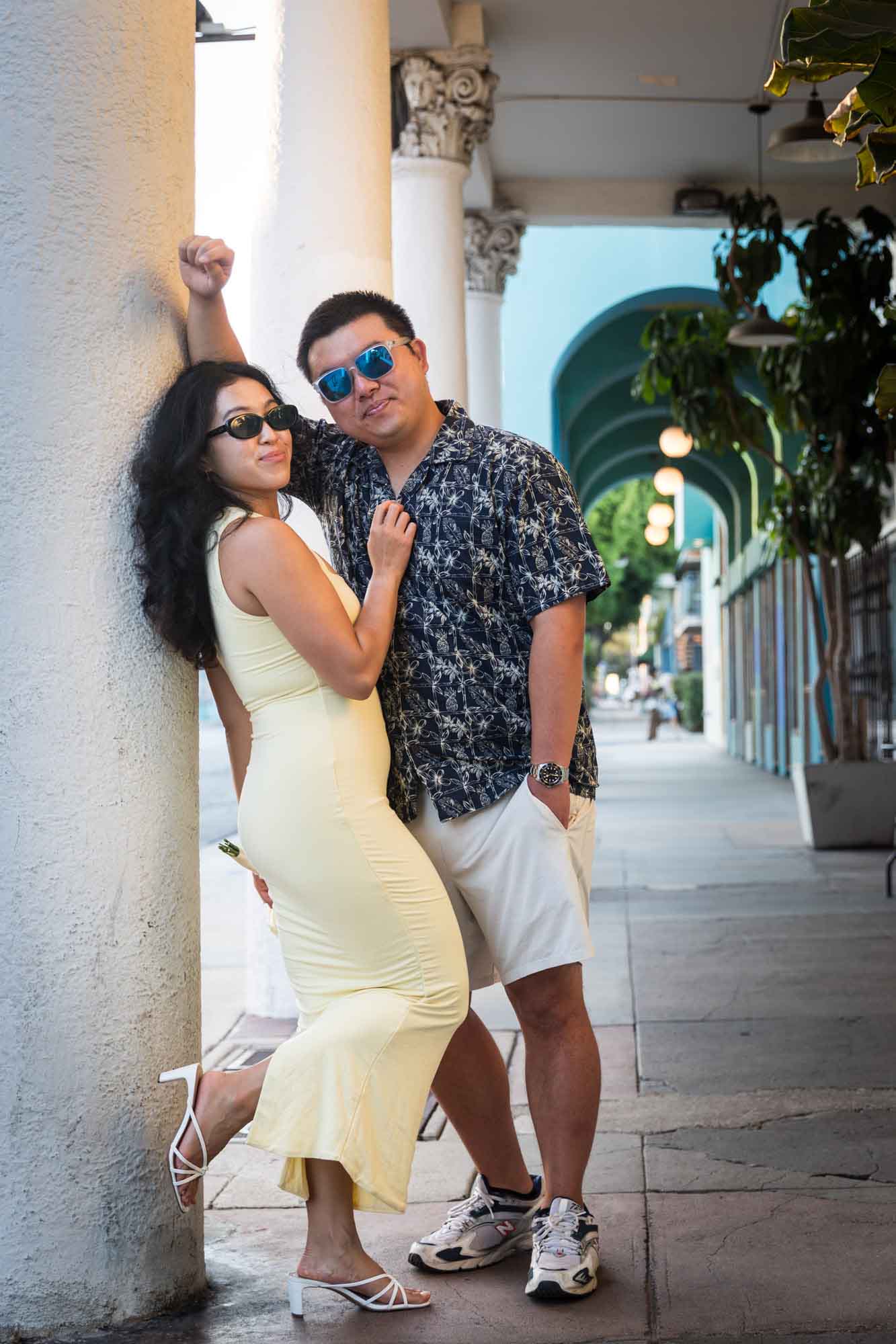 Man wearing blue and white patterned shirt and sunglasses and woman wearing sleeveless yellow dress and sunglasses leaning against column during a Venice Canals engagement photo shoot