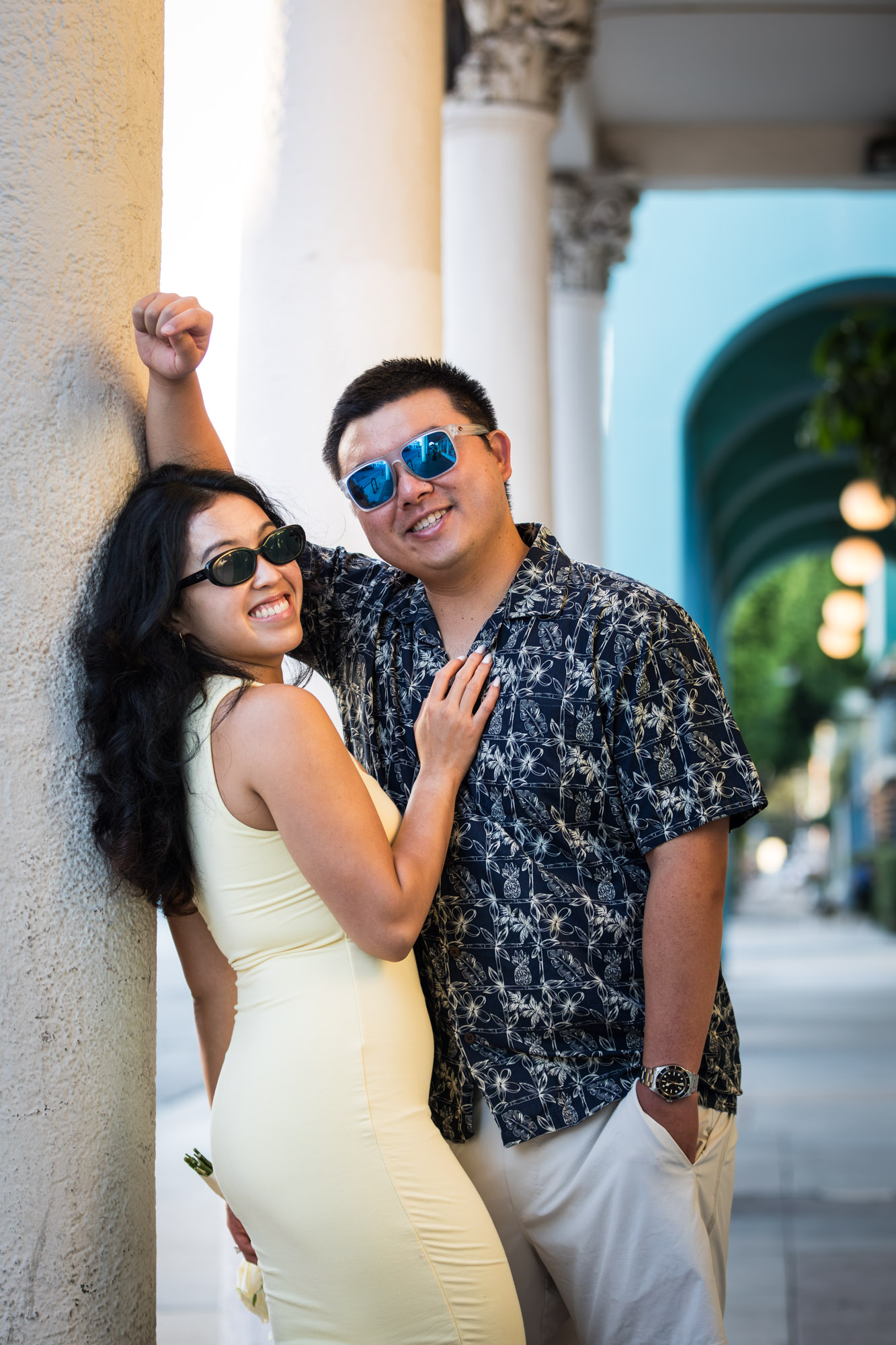 Man wearing blue and white patterned shirt and sunglasses and woman wearing sleeveless yellow dress and sunglasses leaning against column during a Venice Canals engagement photo shoot