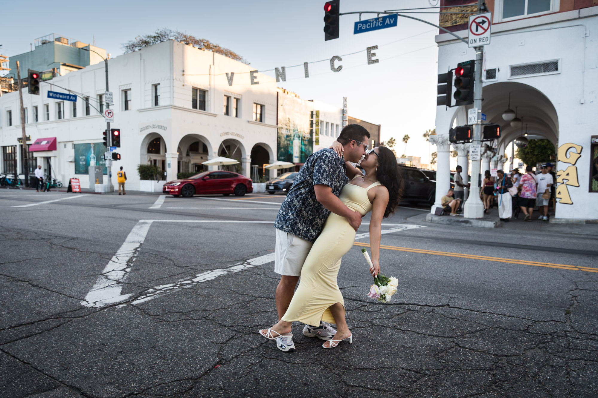 Man wearing blue and white patterned shirt and sunglasses and woman wearing sleeveless yellow dress and sunglasses kissing in the middle of a street intersection with the Venice sign in the background