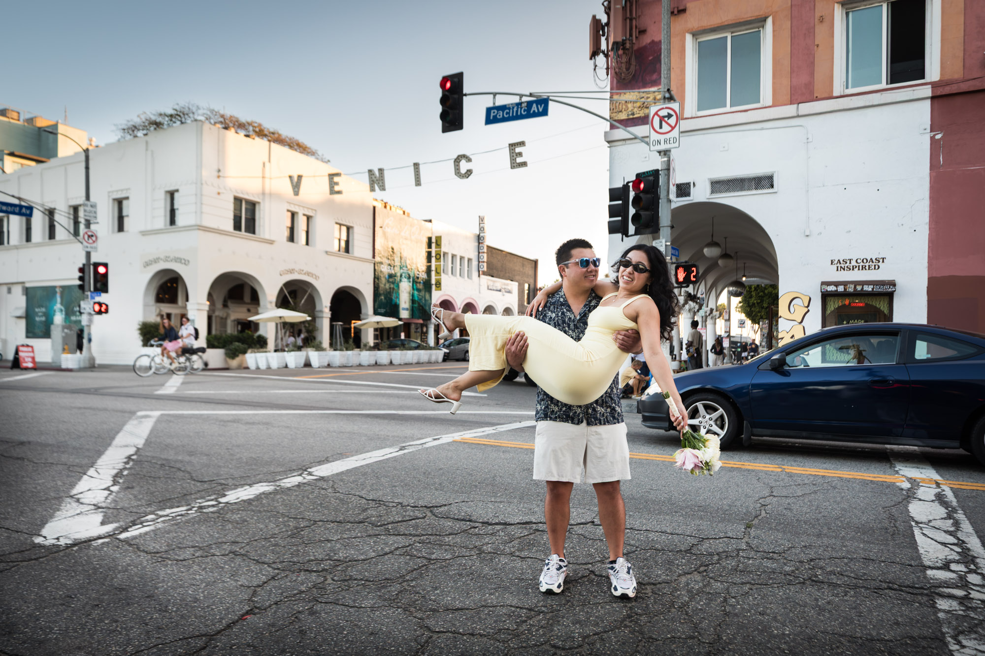 Man wearing blue and white patterned shirt and sunglasses lifting up a woman wearing sleeveless yellow dress and sunglasses in the middle of a street intersection with the Venice sign in the background