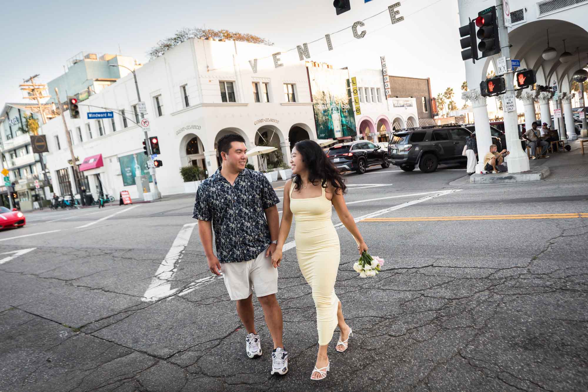 Man wearing blue and white patterned shirt and woman wearing sleeveless yellow dress walking in the middle of a street intersection with the Venice sign in the background