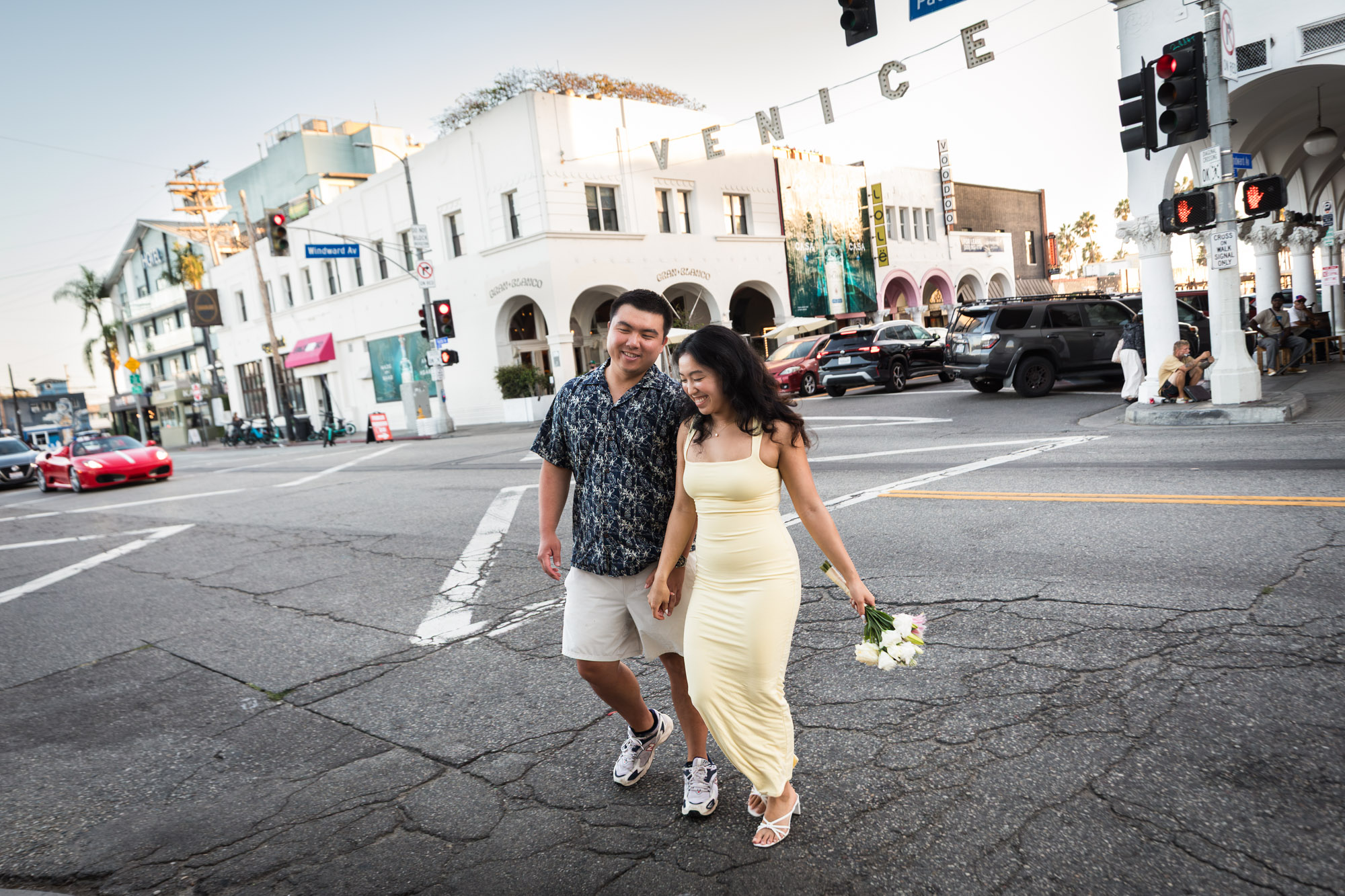 Man wearing blue and white patterned shirt and woman wearing sleeveless yellow dress walking in the middle of a street intersection with the Venice sign in the background