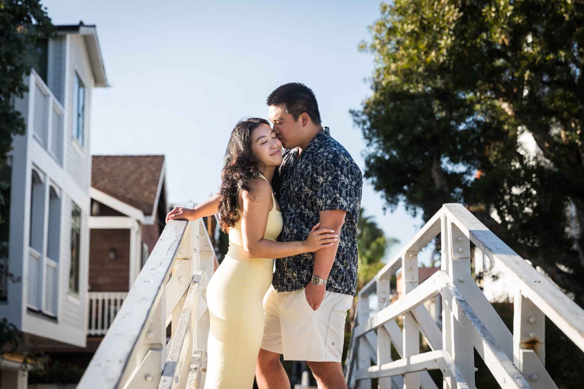 Woman wearing long yellow dress being kissed on the head on a white bridge by man wearing blue and white patterned shirt during a Venice Canals engagement photo shoot