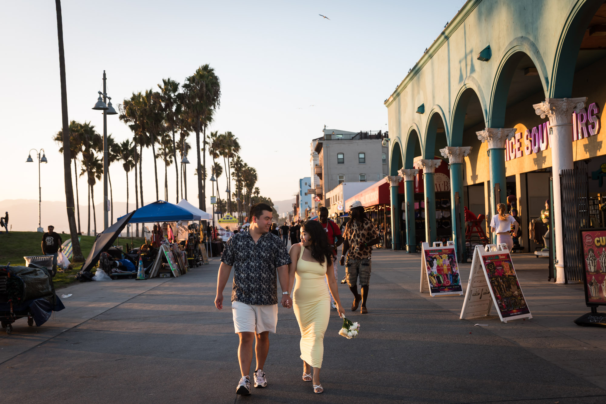 Man wearing blue and white patterned shirt and woman wearing sleeveless yellow dress walking in the middle of the Venice boardwalk with people and A-frame signs surrounding them