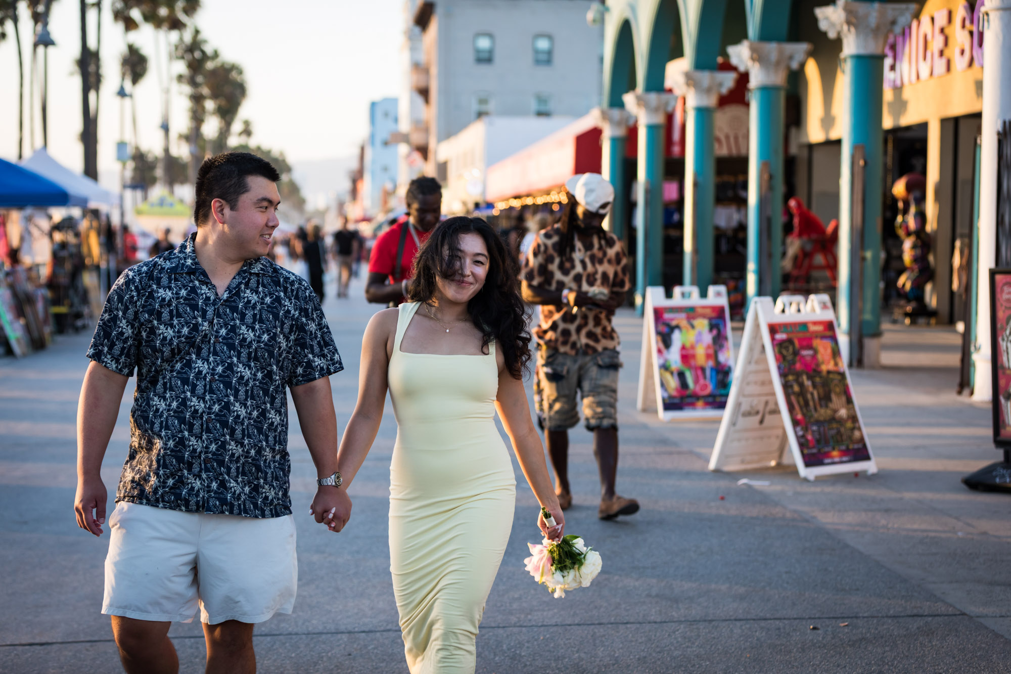 Man wearing blue and white patterned shirt and woman wearing sleeveless yellow dress walking in the middle of the Venice boardwalk with people and A-frame signs surrounding them