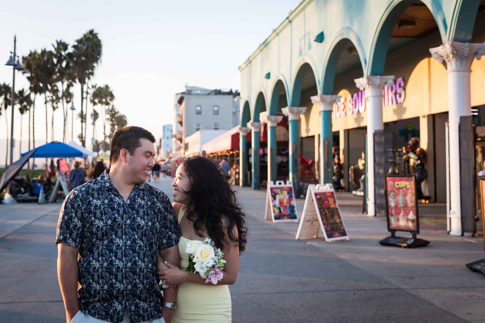 Man wearing blue and white patterned shirt and woman wearing sleeveless yellow dress walking in the middle of the Venice boardwalk with people and A-frame signs surrounding them
