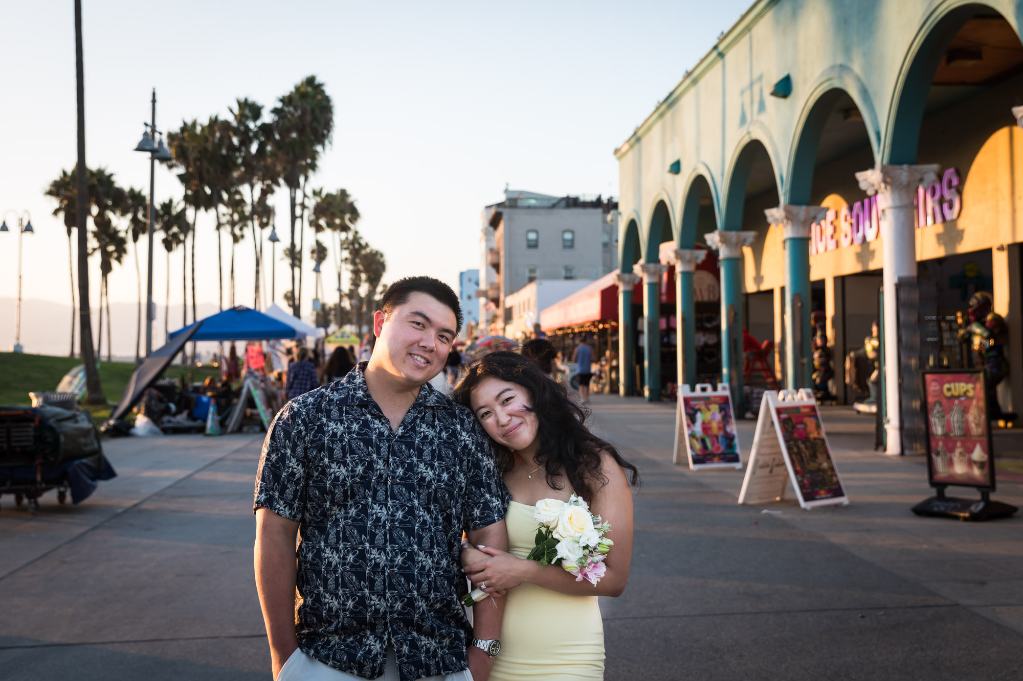 Man wearing blue and white patterned shirt and woman wearing sleeveless yellow dress walking in the middle of the Venice boardwalk with people and A-frame signs surrounding them