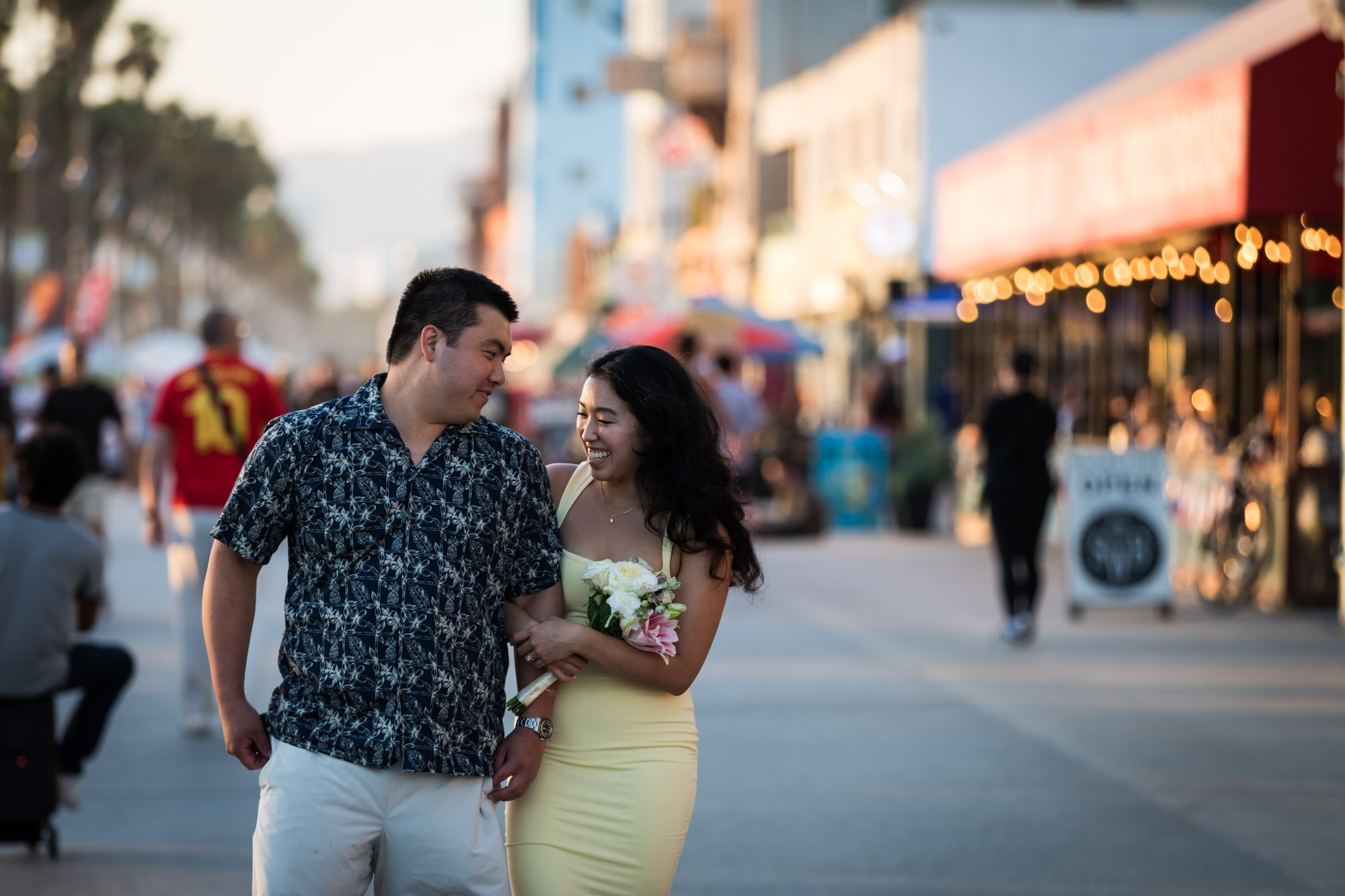 Man wearing blue and white patterned shirt and woman wearing sleeveless yellow dress walking in the middle of the Venice boardwalk with people and A-frame signs surrounding them