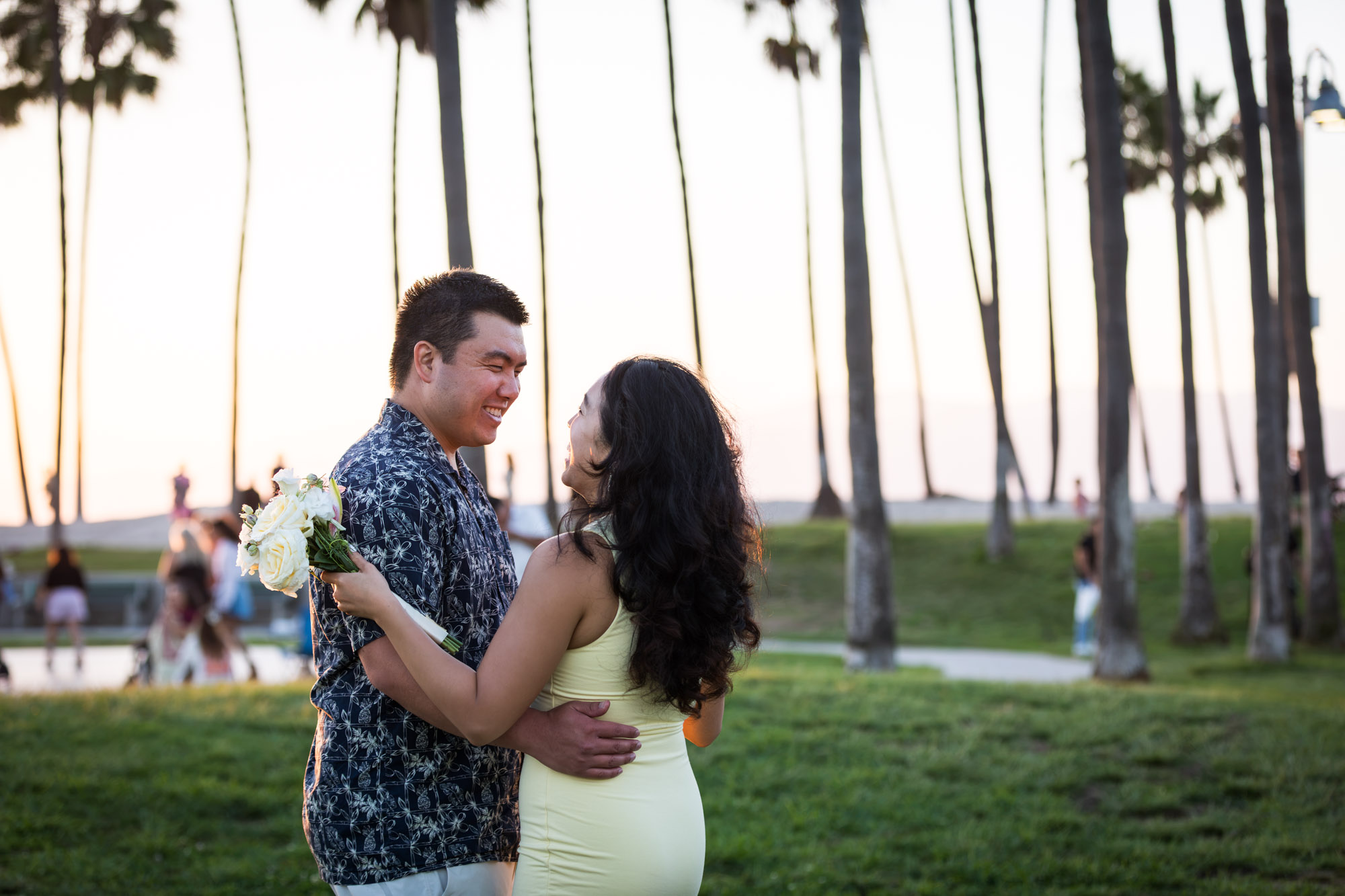 Man wearing blue and white patterned shirt and woman dancing with a woman wearing sleeveless yellow dress in the grass with palm trees in the background at sunset