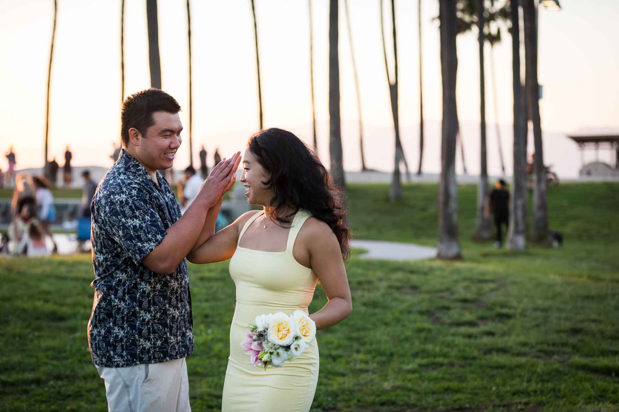 Man wearing blue and white patterned shirt and woman holding hands with a woman wearing sleeveless yellow dress in the grass with palm trees in the background at sunset