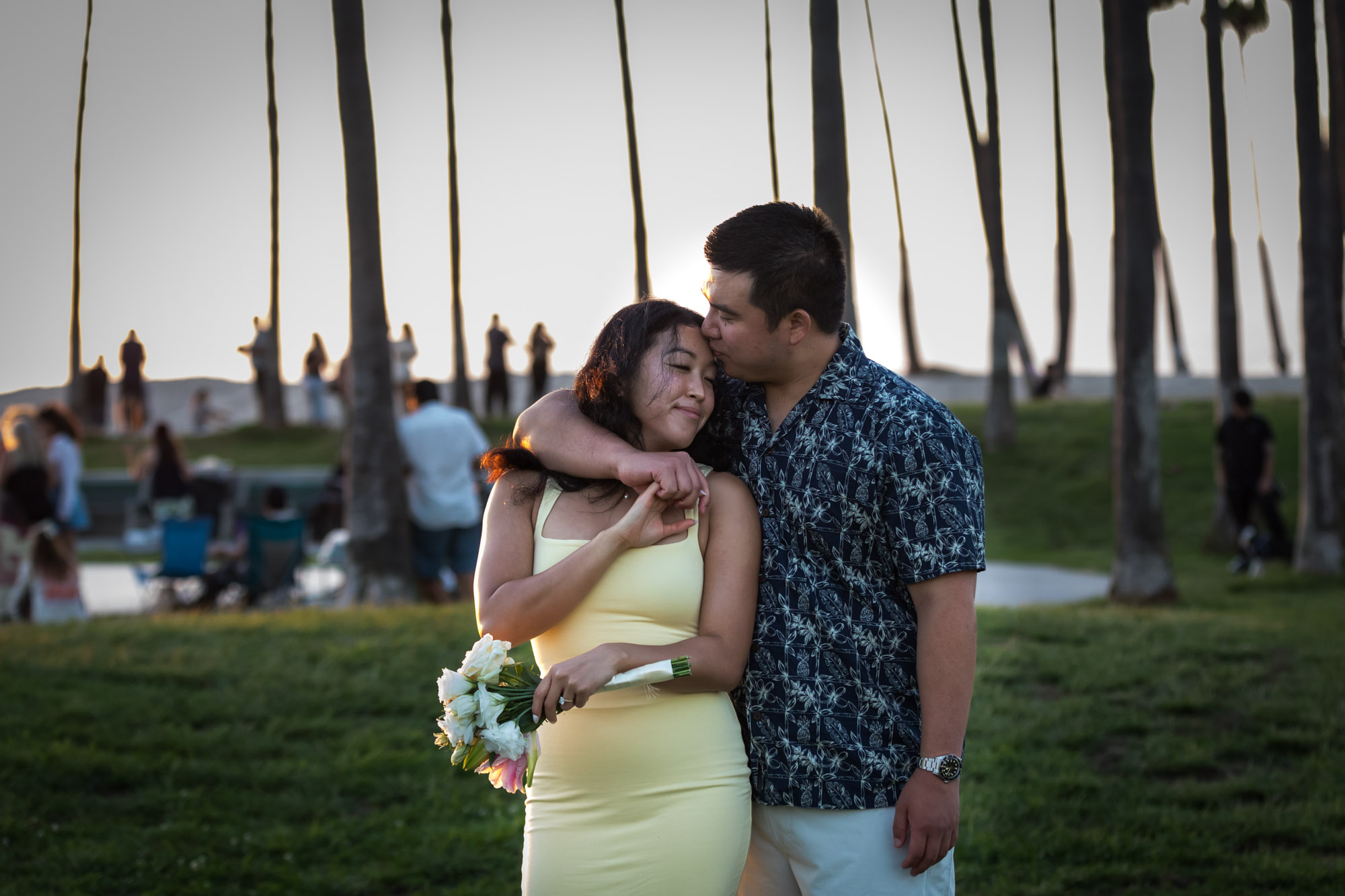 Man wearing blue and white patterned shirt and woman kissing a woman wearing sleeveless yellow dress in the grass with palm trees in the background at sunset