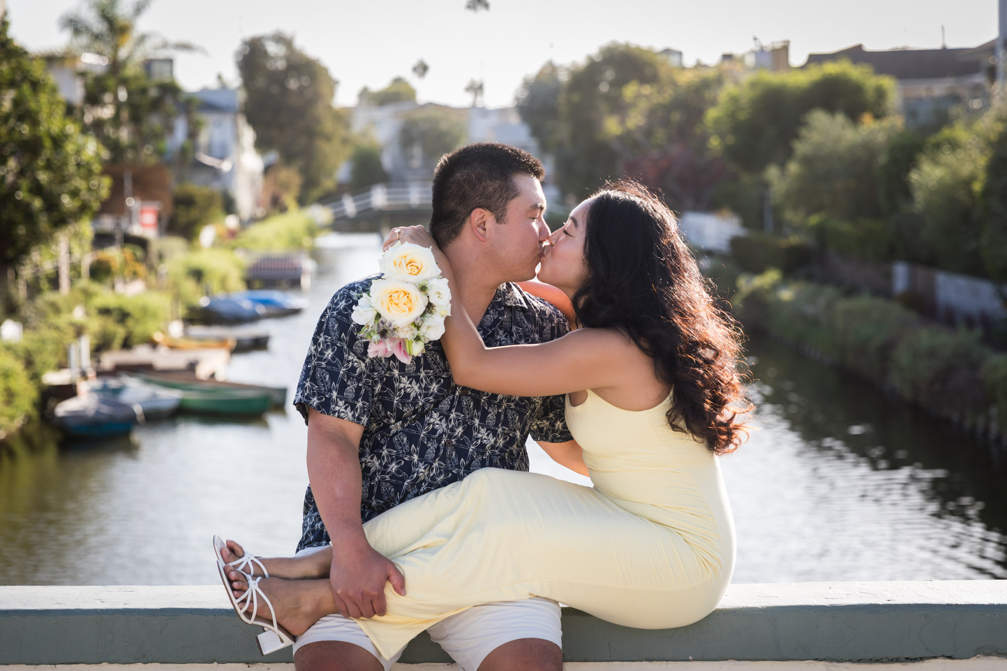 Woman wearing sleeveless yellow dress kissing man wearing blue and white patterned shirt with canals in background during a Venice Canals engagement photo shoot