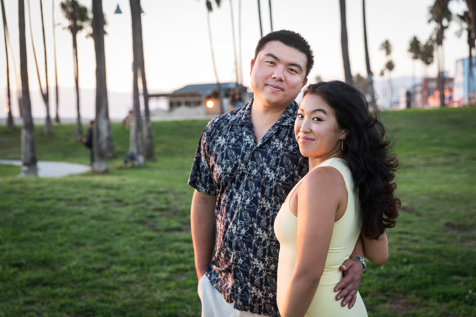 Woman wearing sleeveless, yellow dress and man wearing short-sleeved blue and white shirt standing in grass at sunset with palm trees in background