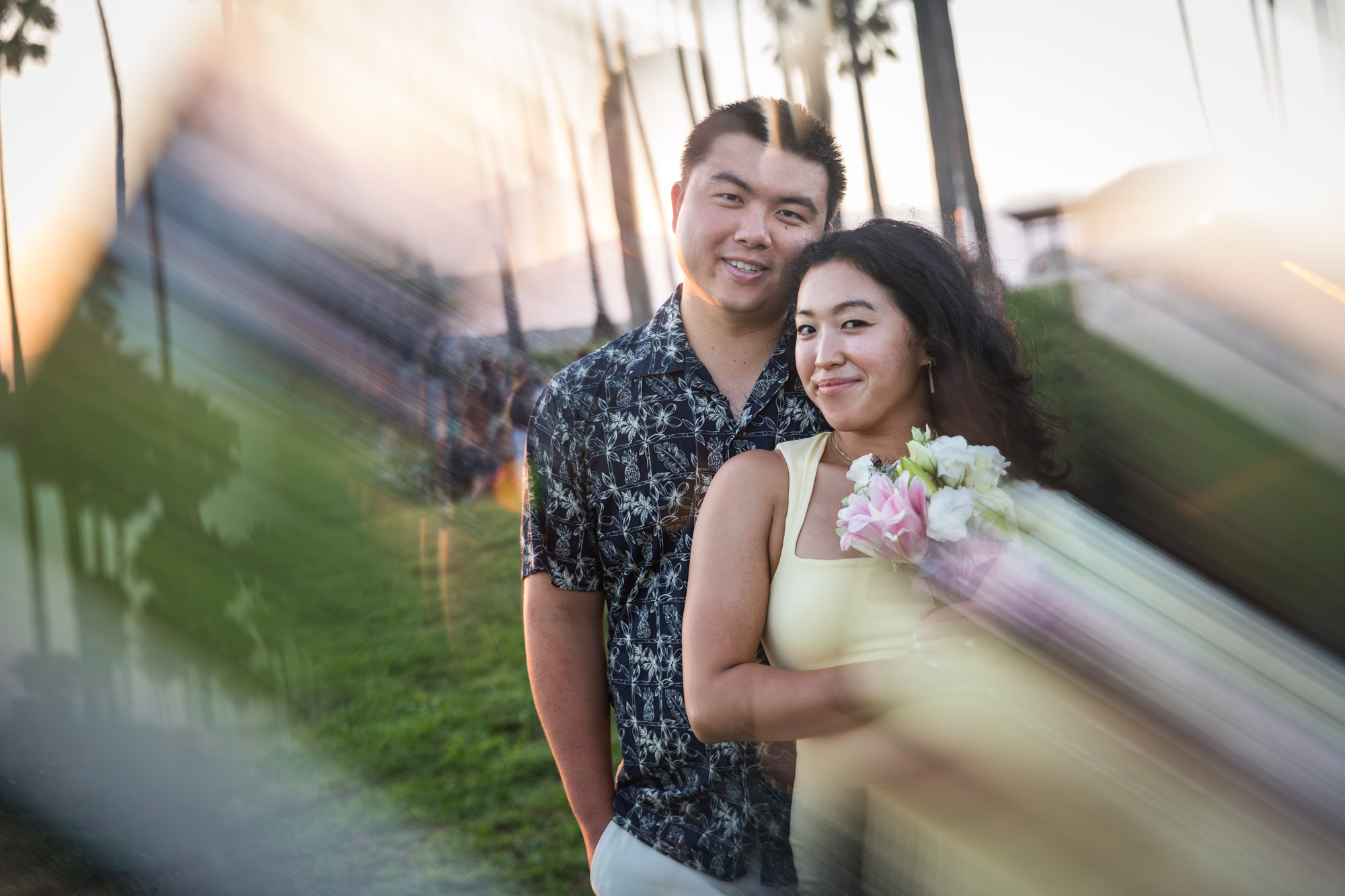 Prism photo of woman wearing sleeveless, yellow dress holding flower bouquet and man wearing short-sleeved blue and white shirt standing in grass at sunset with palm trees in background