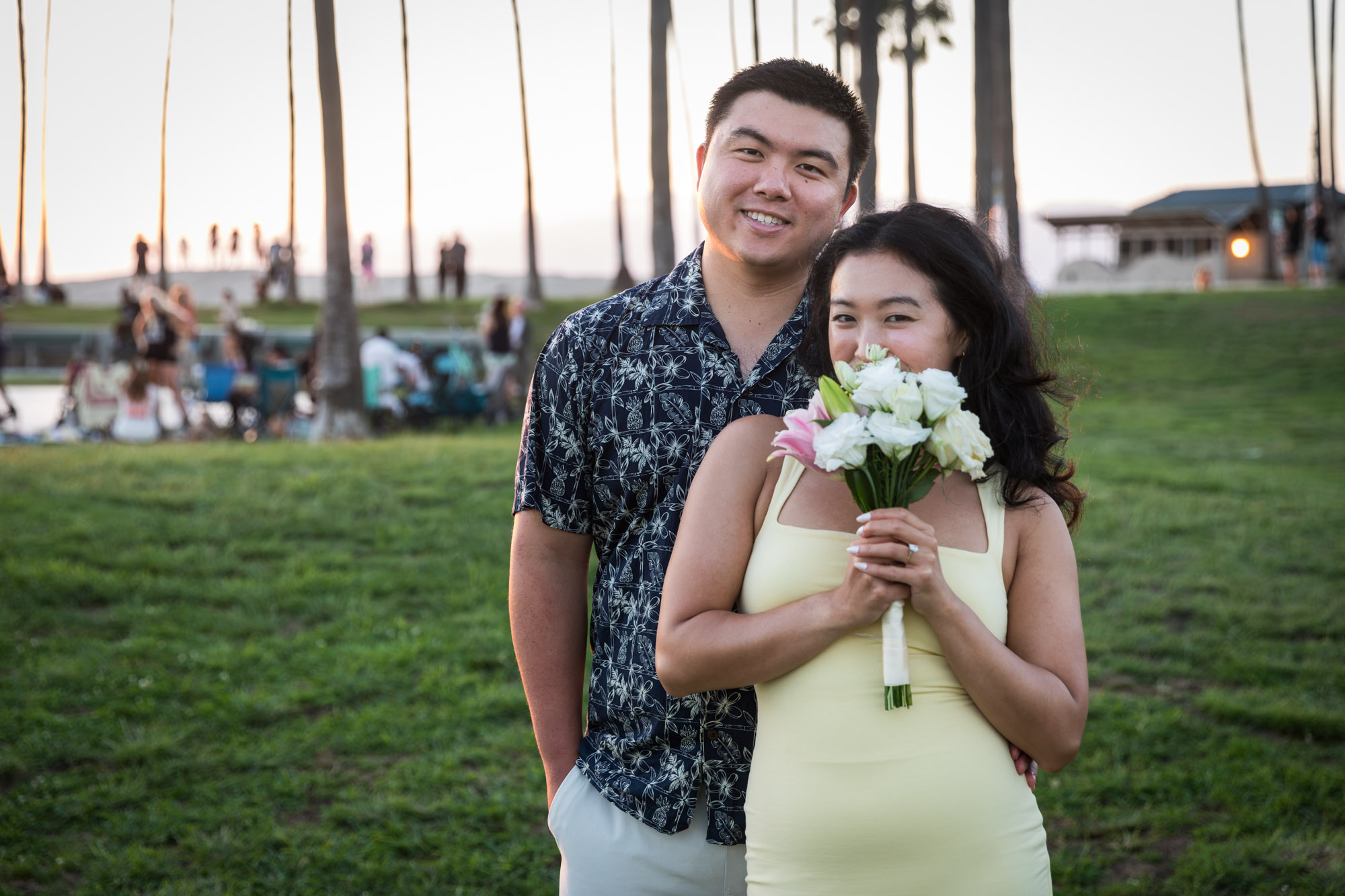 Woman wearing sleeveless, yellow dress holding flower bouquet and man wearing short-sleeved blue and white shirt standing in grass at sunset with palm trees in background