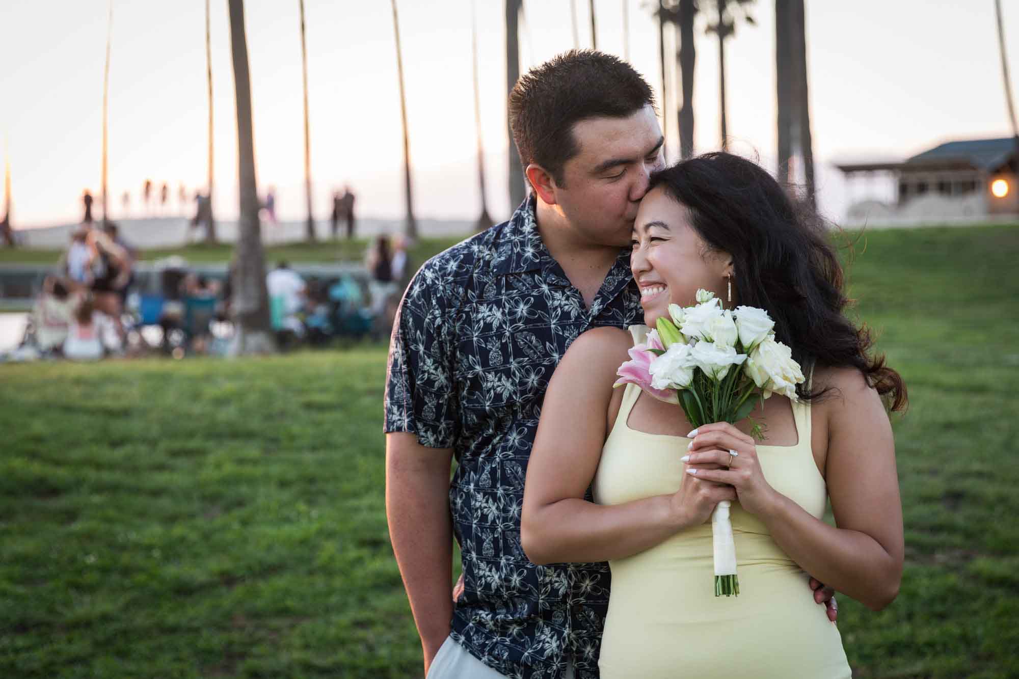 Woman wearing sleeveless, yellow dress being kissed on head by man wearing short-sleeved blue and white shirt standing in grass at sunset with palm trees in background